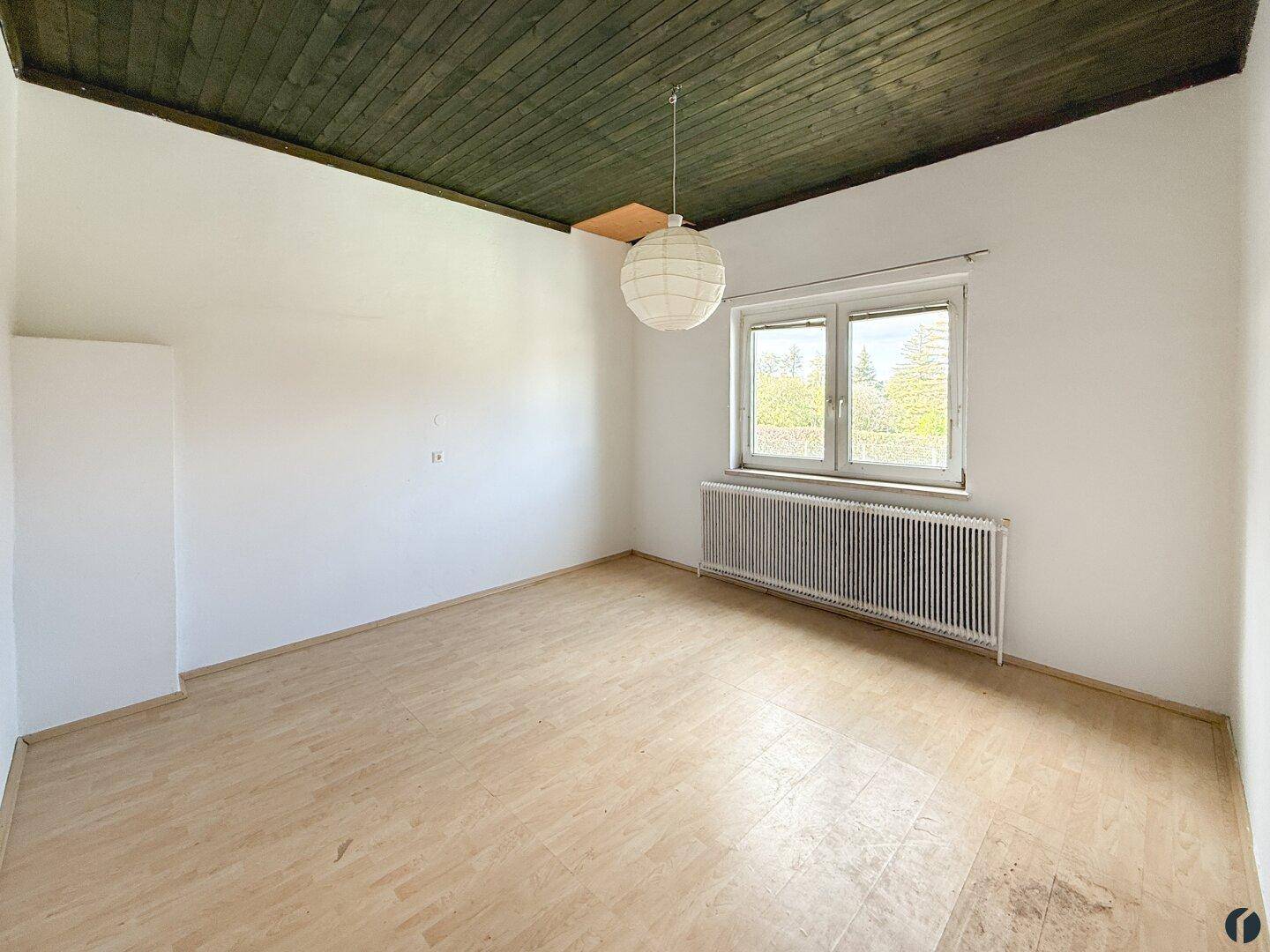Leeres Zimmer mit weißen Wänden, Holzfußboden, Fenster und einer dunklen Holzdecke.