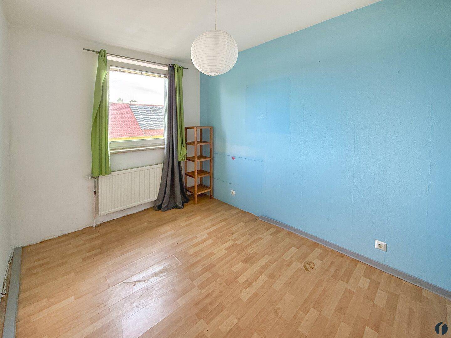 Leeres Zimmer mit blau gestrichener Wand, Holzfußboden und einem Fenster mit grünen Vorhängen.