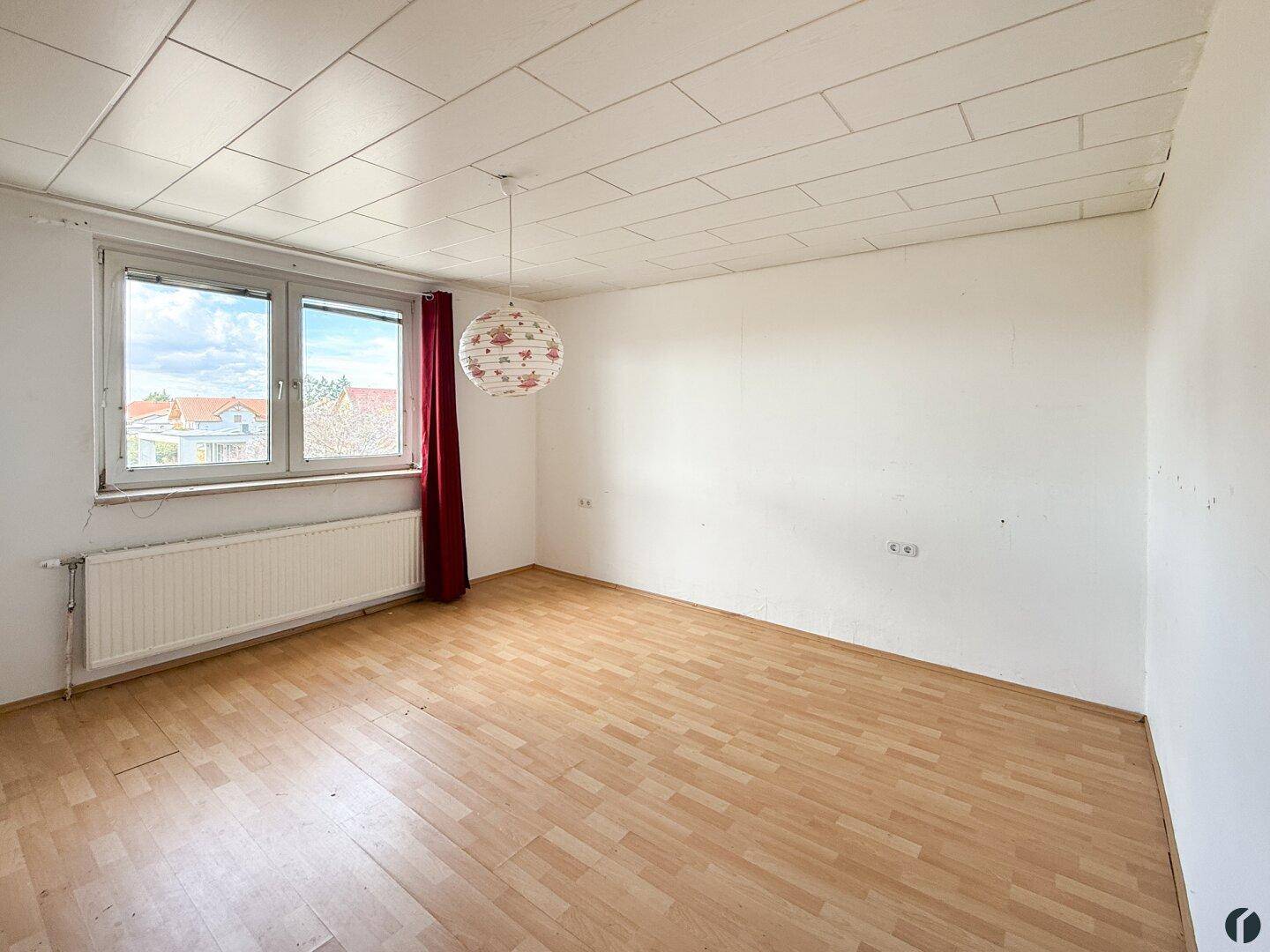 Helles Zimmer mit Holzfußboden, weißen Wänden, Fenster mit roten Vorhängen und einer weißen Decke.