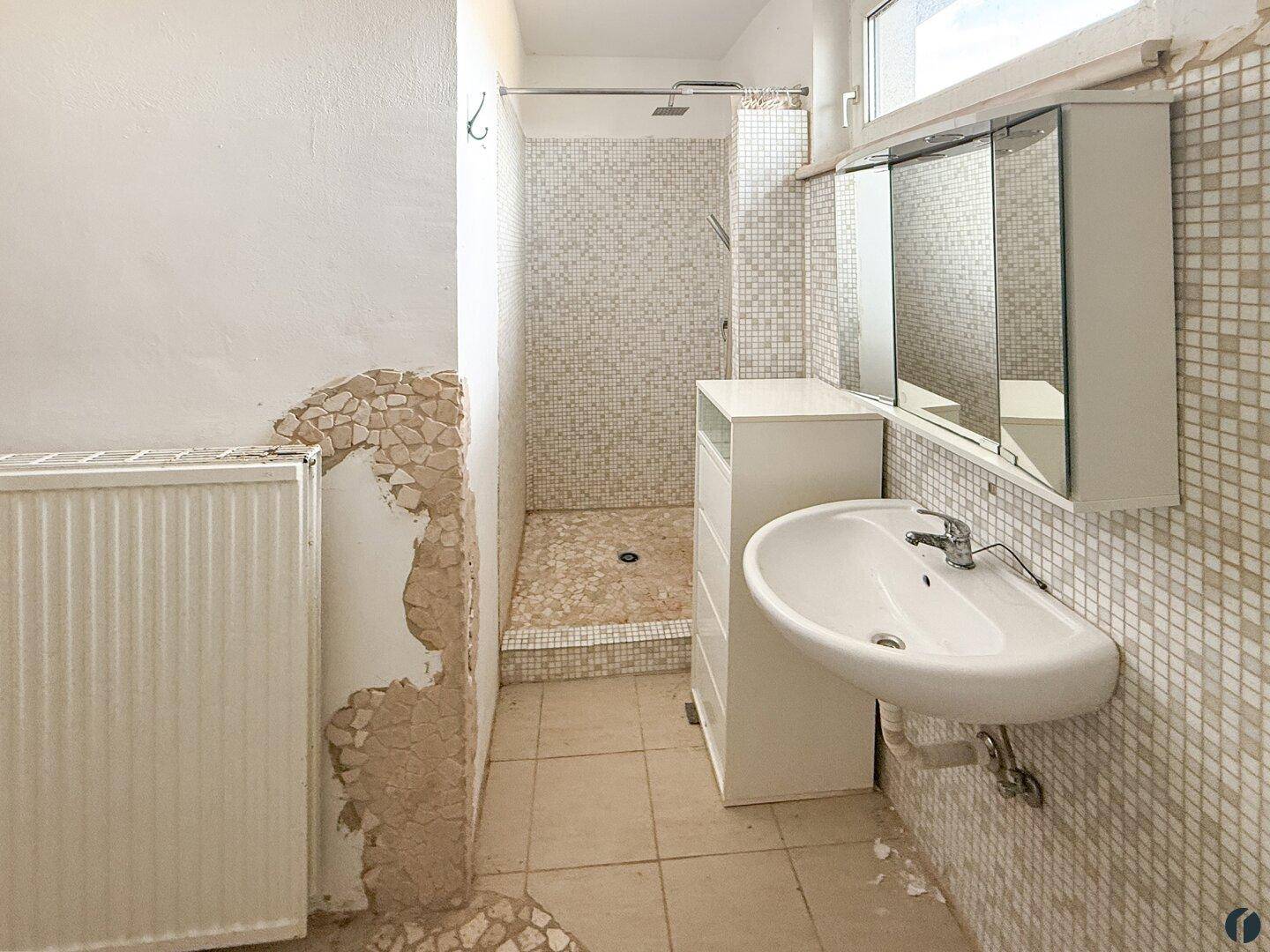 Badezimmer mit Dusche, Waschbecken, Spiegelschrank und Fliesenboden, renovierungsbedürftig.