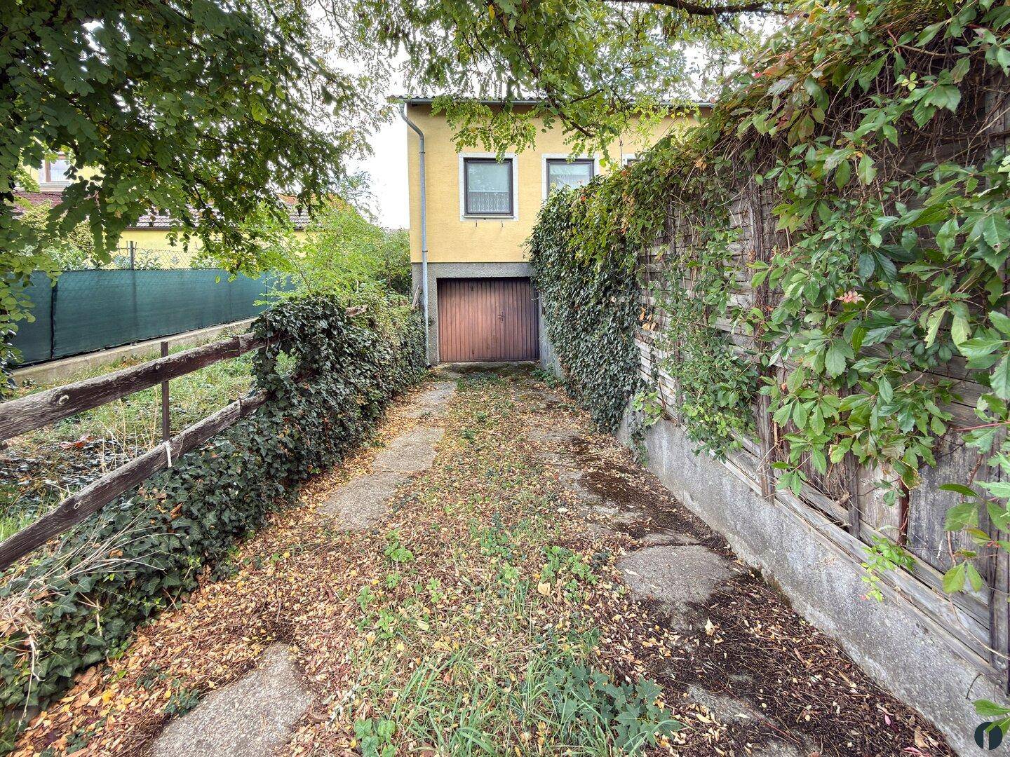 Einfahrt zu einer Garage, umgeben von üppigem Grün und einem Holzzaun.