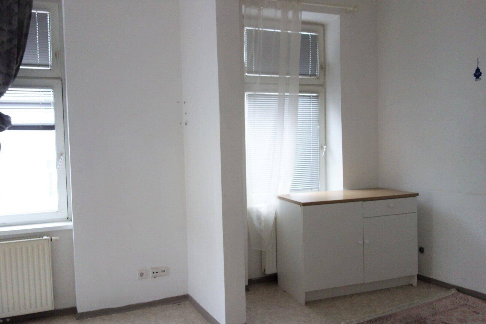 Helles Zimmer mit einem kleinen Schrank und Fenstern, die für natürliche Belichtung sorgen.