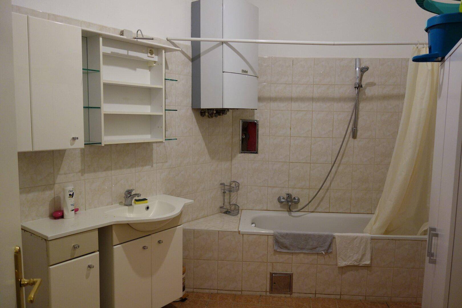Funktionales Badezimmer mit Badewanne, Waschbecken und Warmwasserboiler, gefliest in hellem Beige.