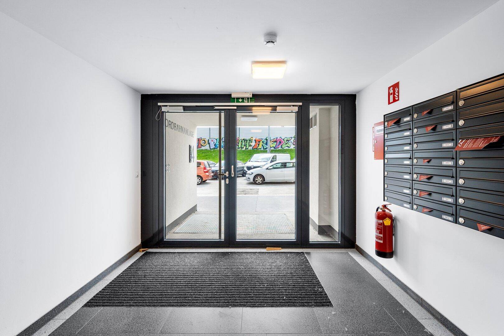Eingangsbereich eines Wohnhauses mit Briefkästen, Feuerlöscher und Blick nach draußen.