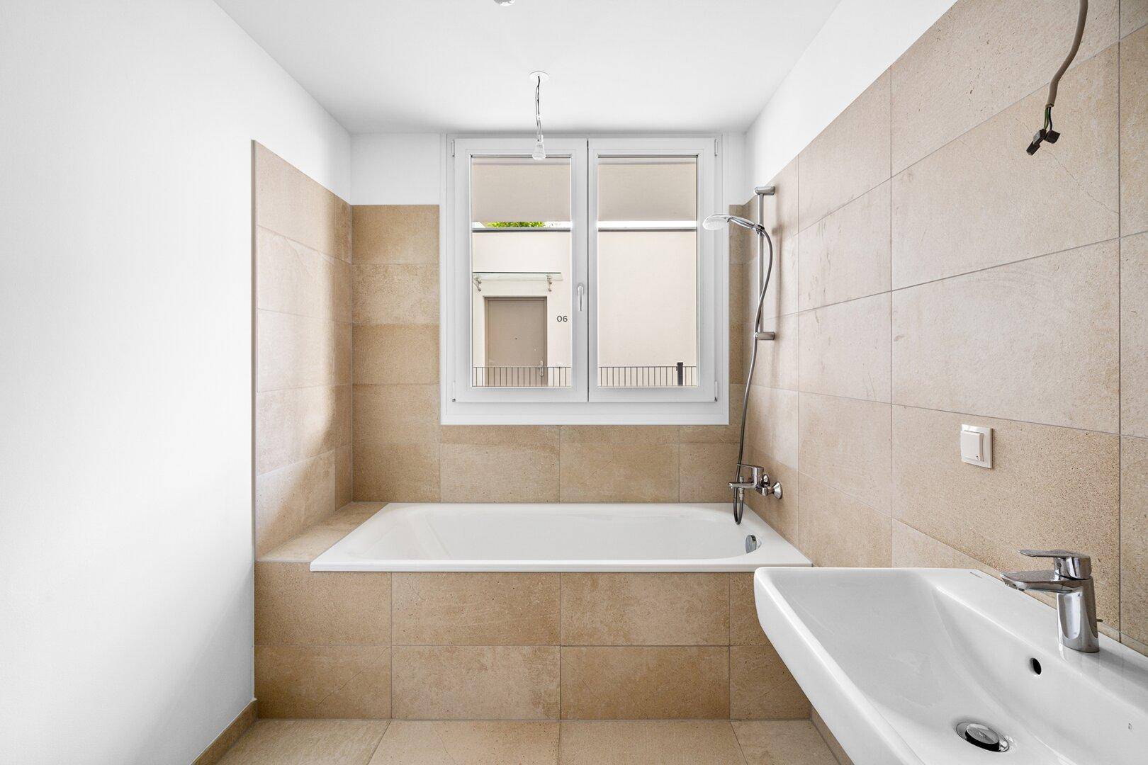 Modernes Badezimmer mit Badewanne, Fenster und hellen Fliesen an Wänden und Boden.