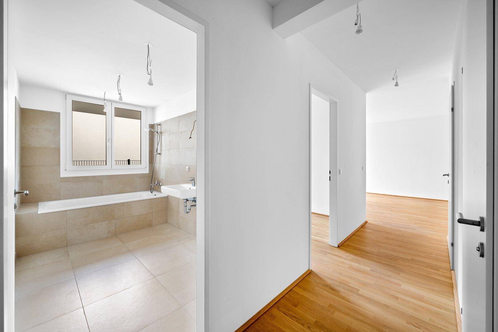 Heller Flur mit Parkettboden und Blick in ein Badezimmer mit Badewanne und Fenster.