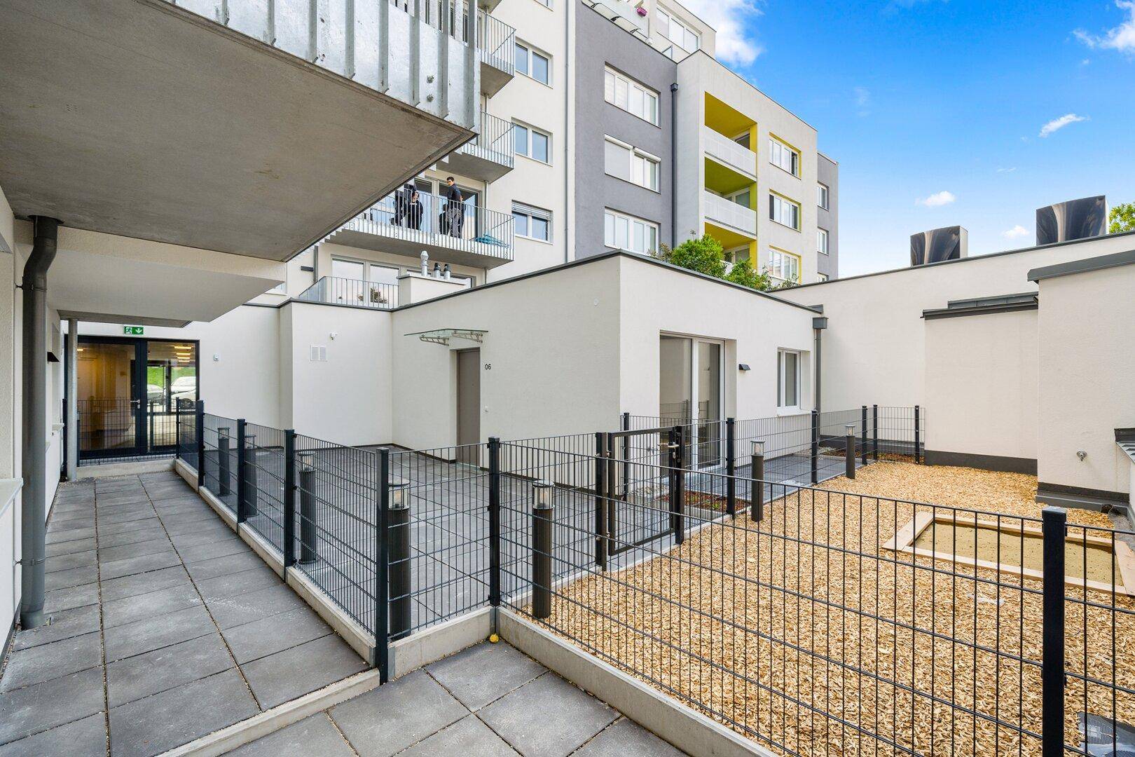 Außenbereich mit gepflastertem Weg und umzäuntem Kiesgarten, umgeben von modernen Gebäuden.