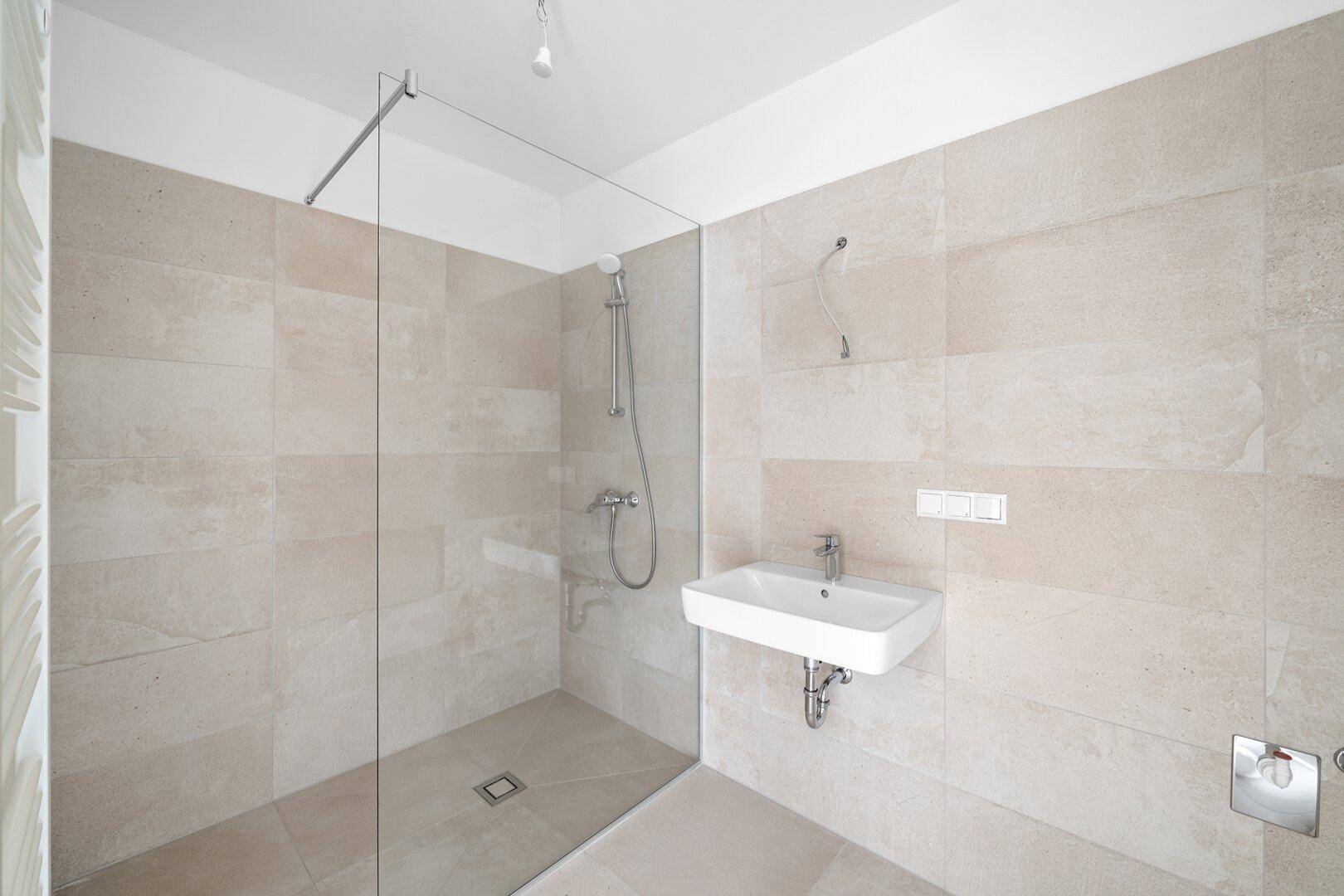 Modernes Badezimmer mit ebenerdiger Dusche, Waschbecken und hellen Fliesen an Wänden und Boden.