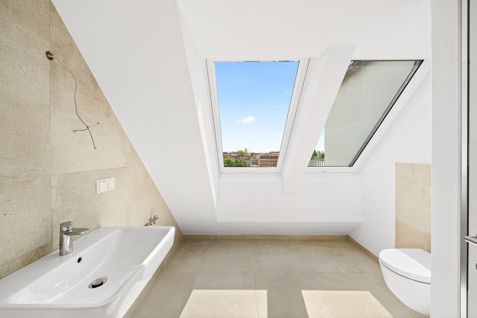 Modernes Badezimmer im Dachgeschoss mit Dachfenster, Waschbecken und Wand-WC, hell gefliest.