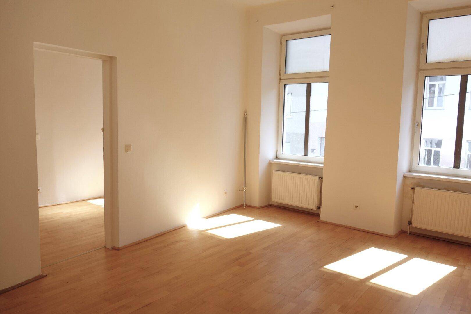 Helles, leeres Zimmer mit Holzboden und zwei großen Fenstern, durch die Sonnenlicht fällt.