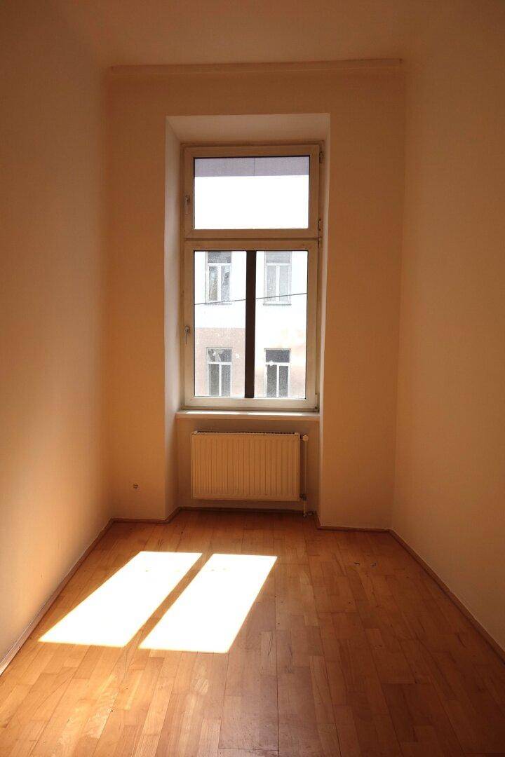 Kleineres, helles Zimmer mit Holzboden und einem Fenster, das Sonnenlicht auf den Boden wirft.