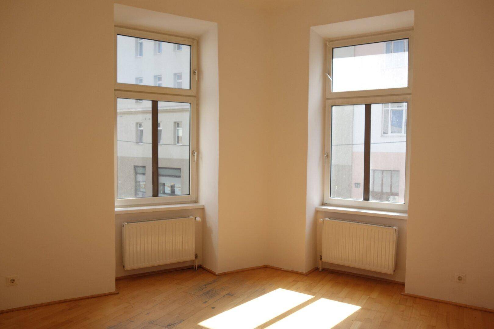 Leeres Zimmer mit Holzboden und zwei Fenstern, die viel Tageslicht hereinlassen.