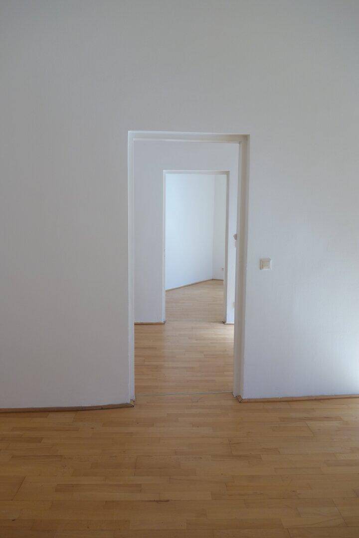 Leerer Raum mit Holzboden und einem offenen Durchgang, der zu weiteren Zimmern führt.