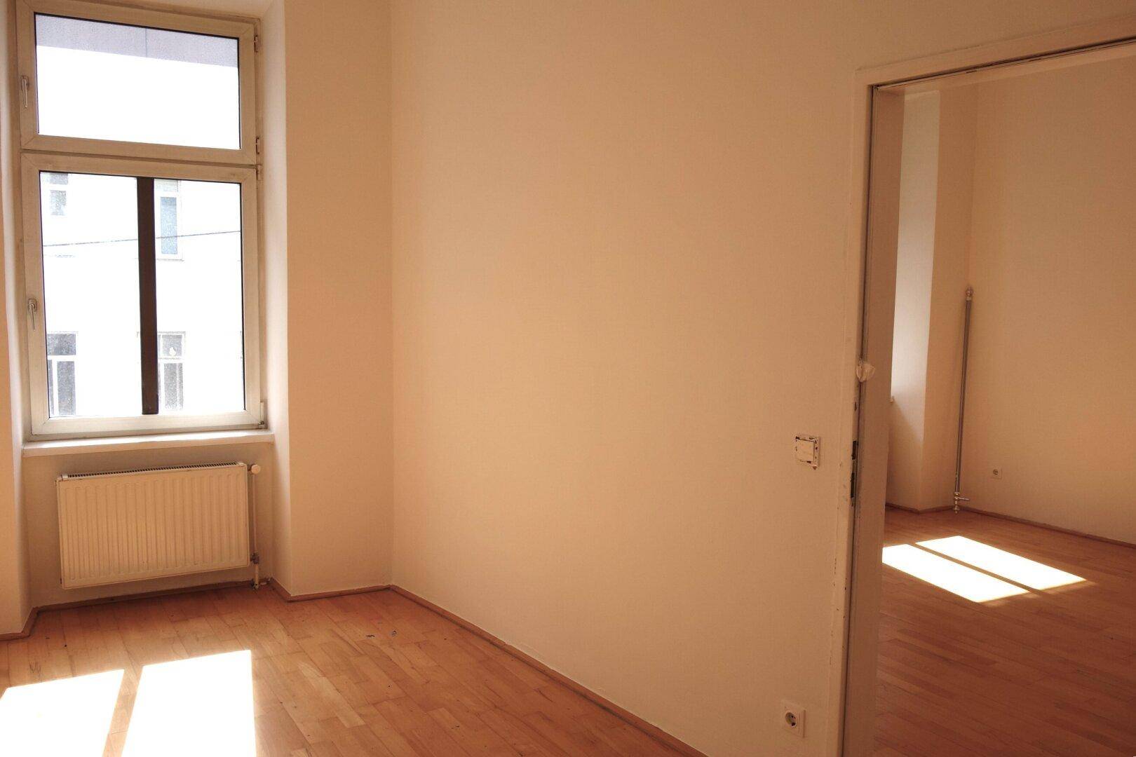 Helles Zimmer mit Holzboden, einem Fenster und einem offenen Durchgang zu einem weiteren Raum.