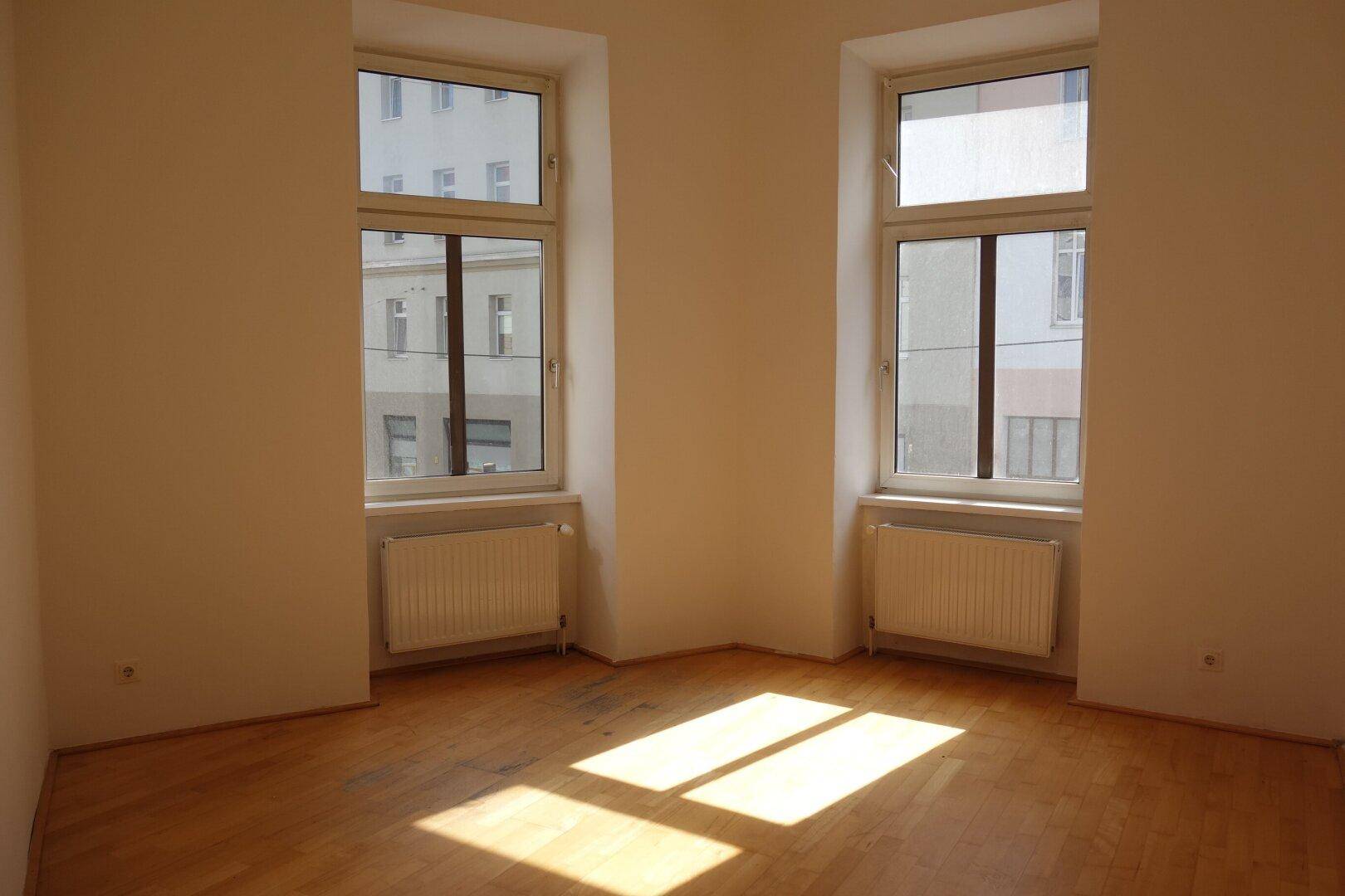 Helles Zimmer mit Holzboden und zwei Fenstern, durch die Sonnenlicht auf den Boden fällt.