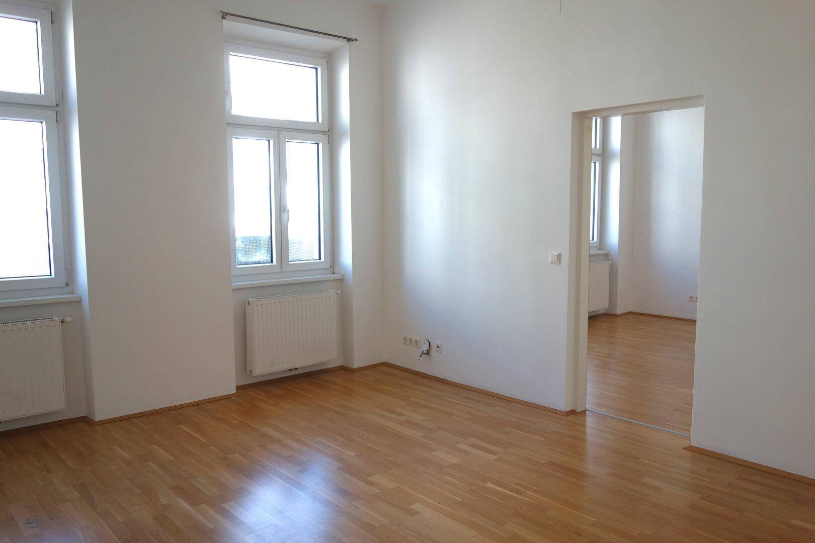 Geräumiges, helles Zimmer mit Holzparkett und zwei großen Fenstern, ideal für Wohnzwecke.