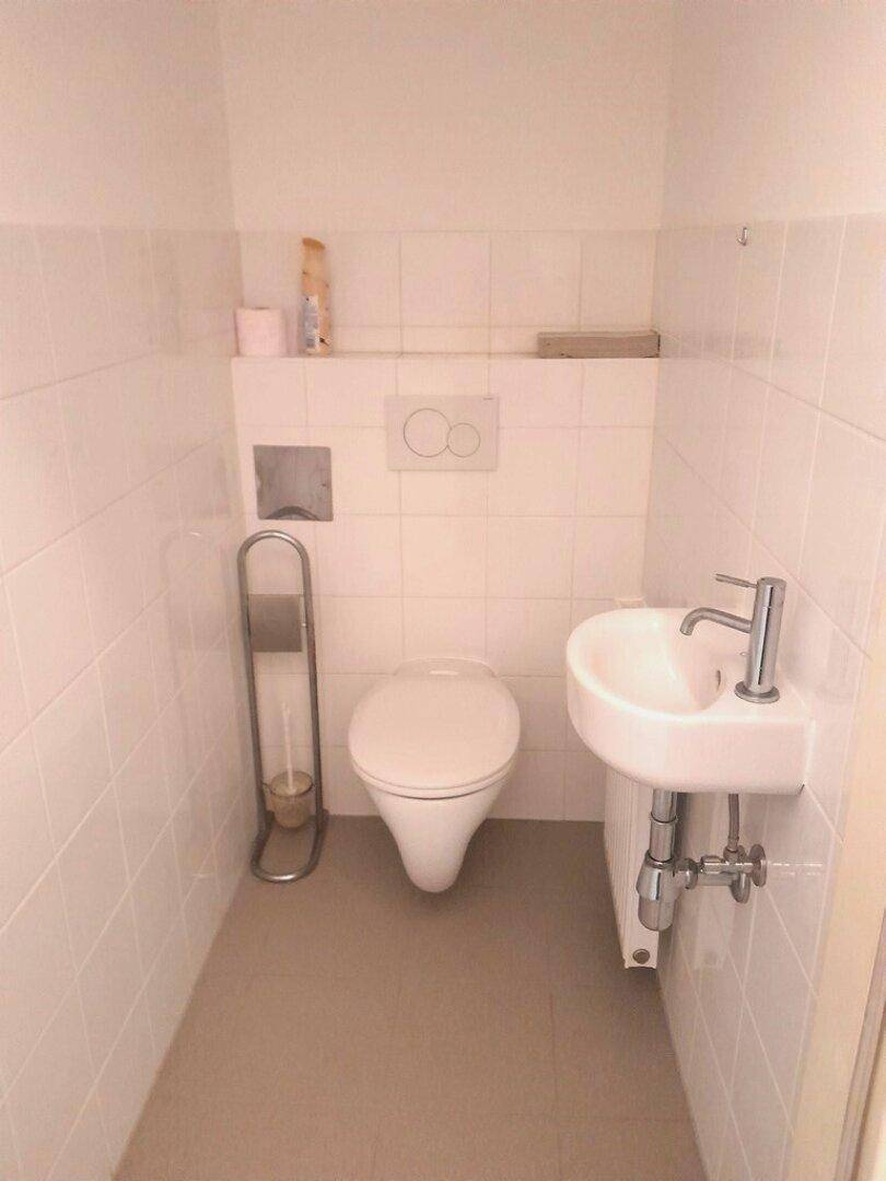 Separates WC mit wandhängender Toilette, kleinem Waschbecken und weißen Fliesen.