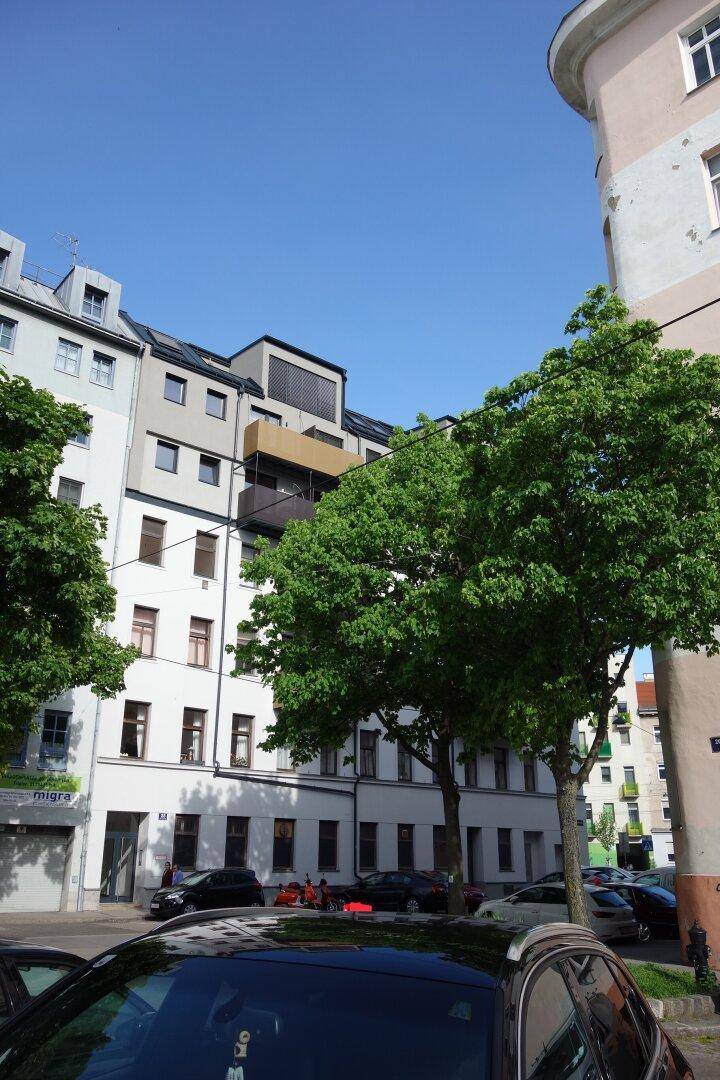 Städtisches Wohngebäude mit Balkonen und Bäumen im Vordergrund unter blauem Himmel.