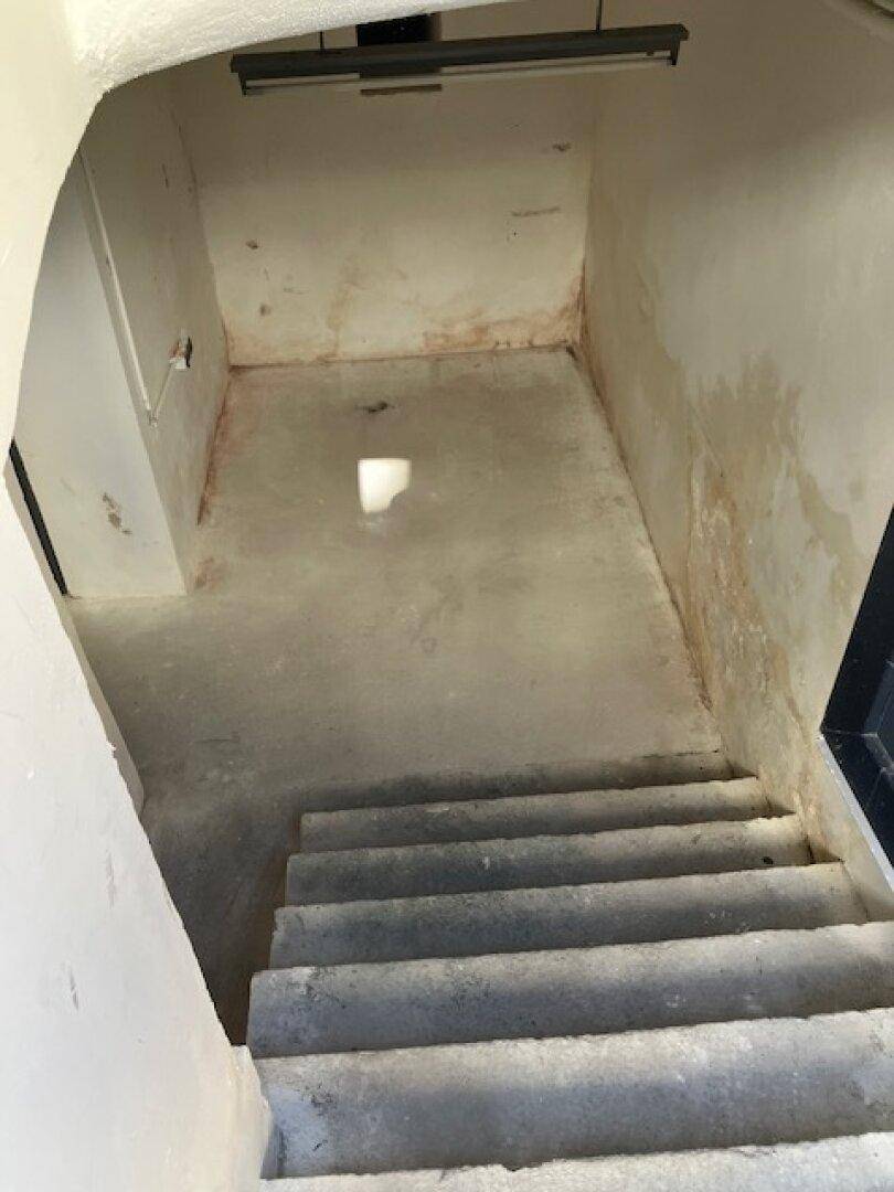 Betontreppe führt hinunter in einen kleinen Kellerraum mit einfacher Beleuchtung und abgenutzten Wänden.