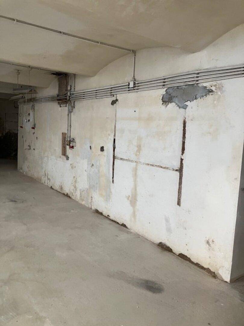 Weiterer Blick in einen Kellerraum mit Betonboden, sichtbaren Leitungen und stark abgenutzten Wänden.