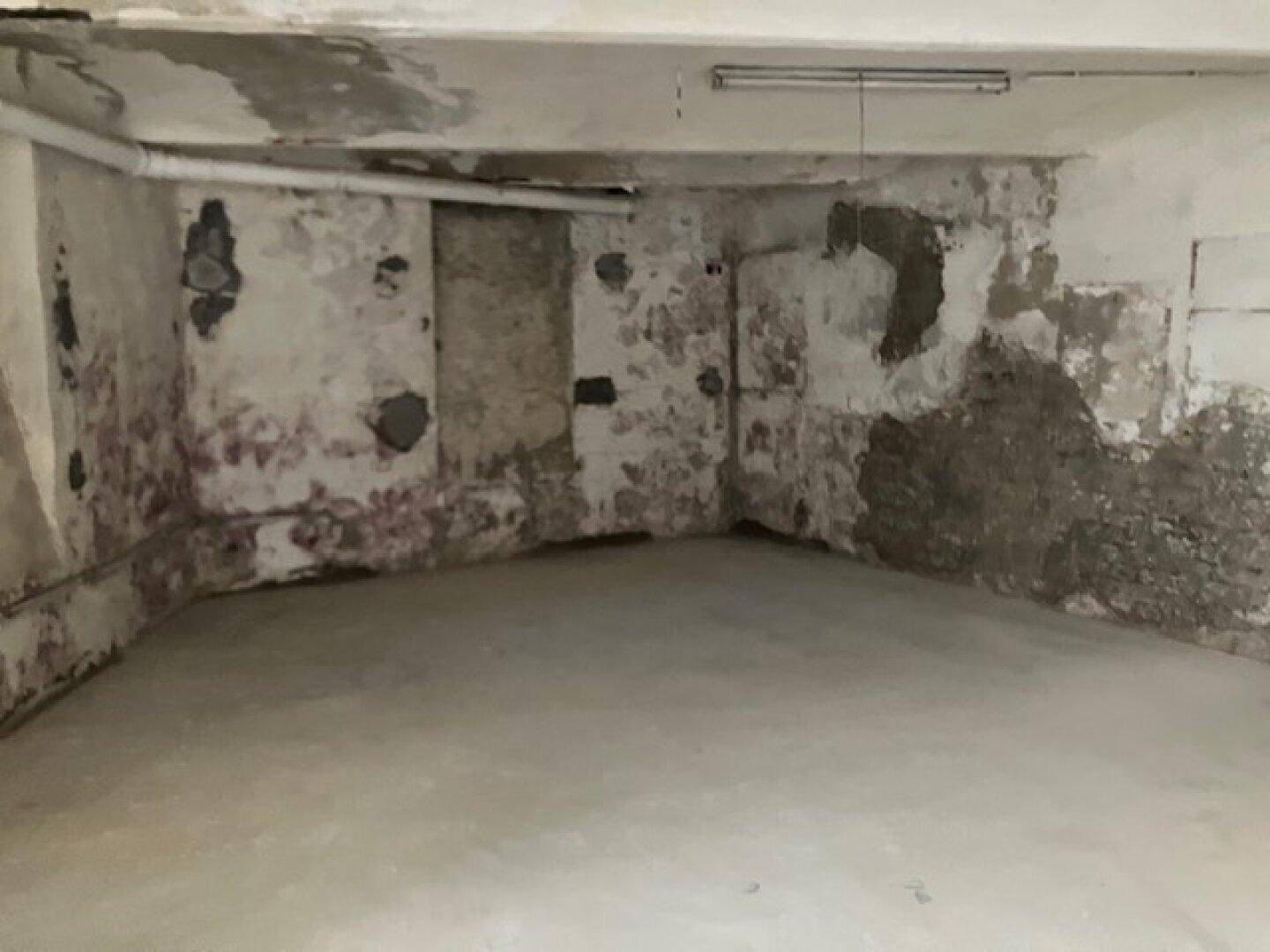 Kellerecke mit Betonboden und stark beschädigten Wänden, die Feuchtigkeit und Ziegelmauerwerk zeigen.