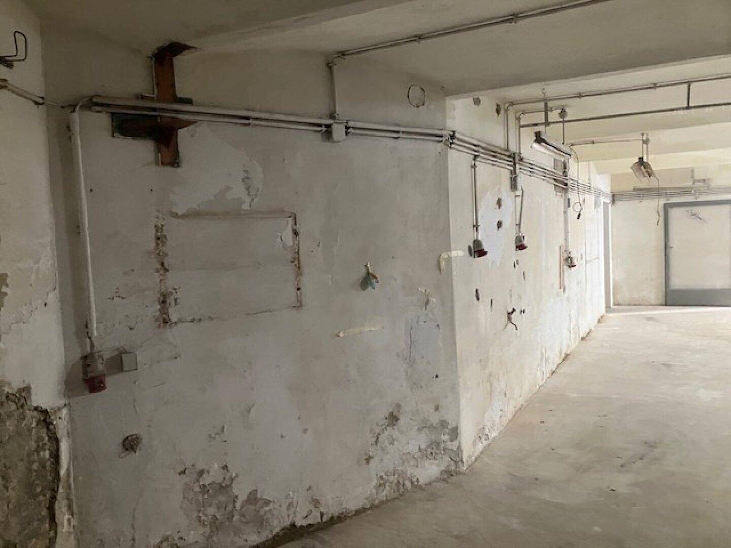 Weiterer Blick in den Kellerraum mit Betonboden, abblätternder Farbe und freiliegenden Installationen.