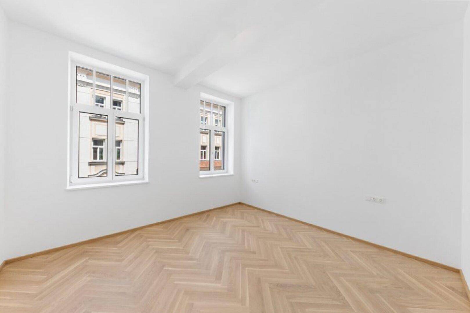 Geräumiges Schlafzimmer mit Fischgrätparkett und viel Tageslicht durch zwei Fenster.