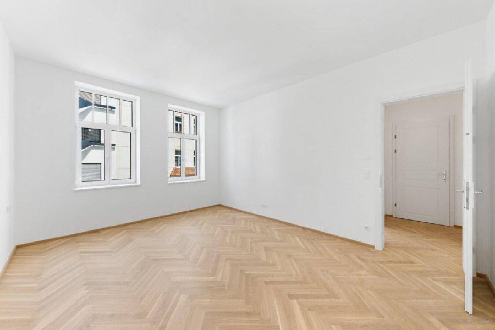 Helles Schlafzimmer mit klassischem Fischgrätparkett und zwei großen Fenstern.