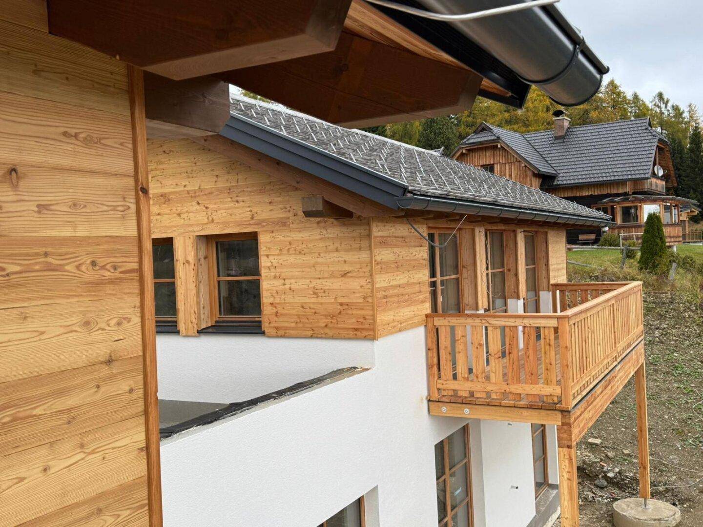 Neubau eines Hauses mit Holzfassade, weißem Putz und einem Holzbalkon, umgeben von Bäumen.