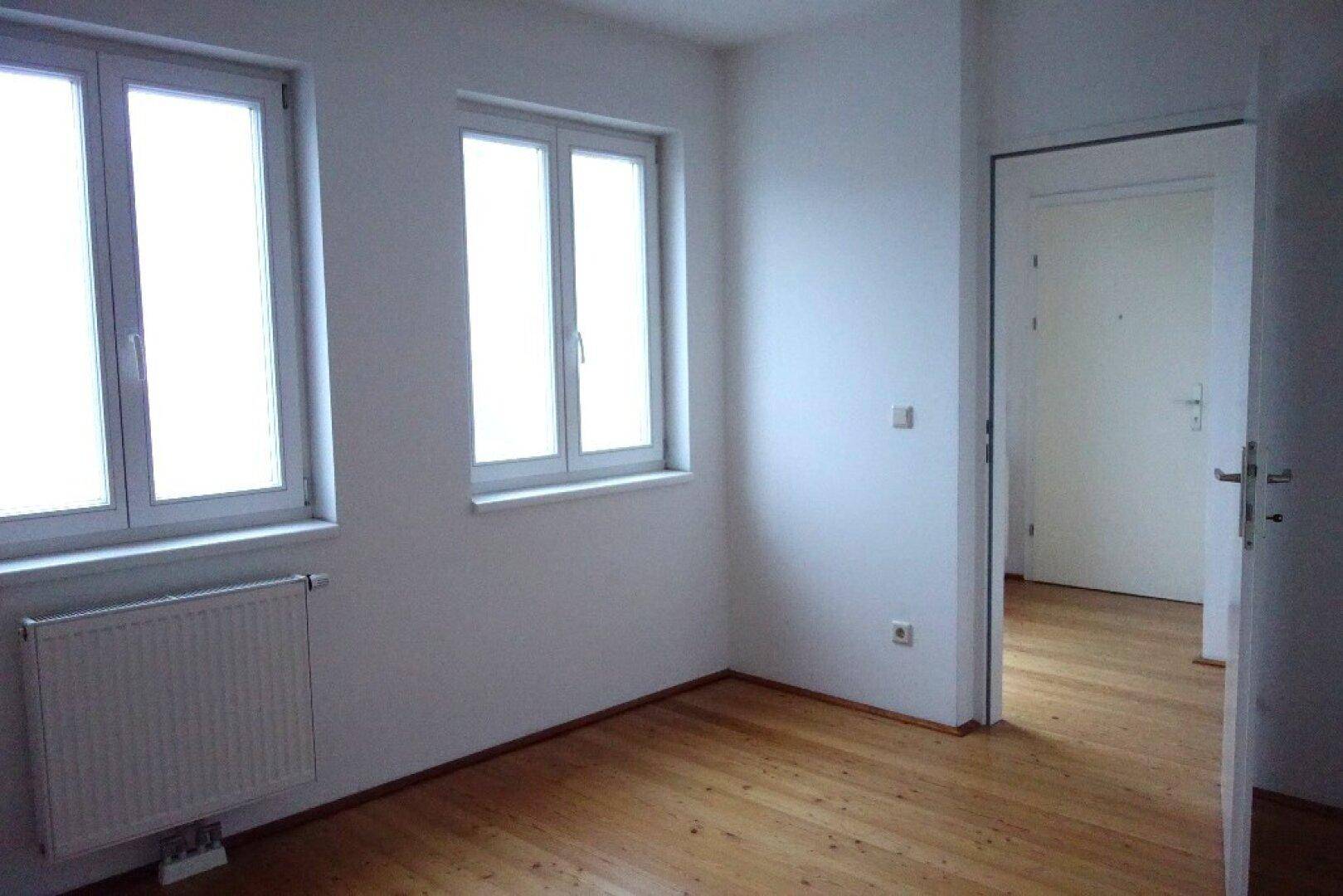 Ruhiges Zimmer mit Holzfußboden und zwei Fenstern, geeignet als Schlaf- oder Arbeitszimmer.