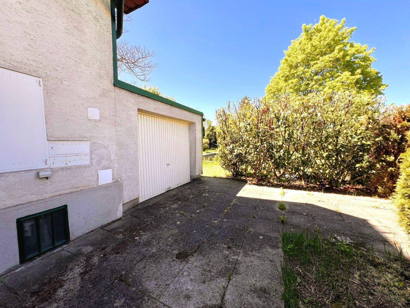 Einzelgarage mit Zufahrt und angrenzender Hecke auf dem Grundstück, beleuchtet von der Sonne.