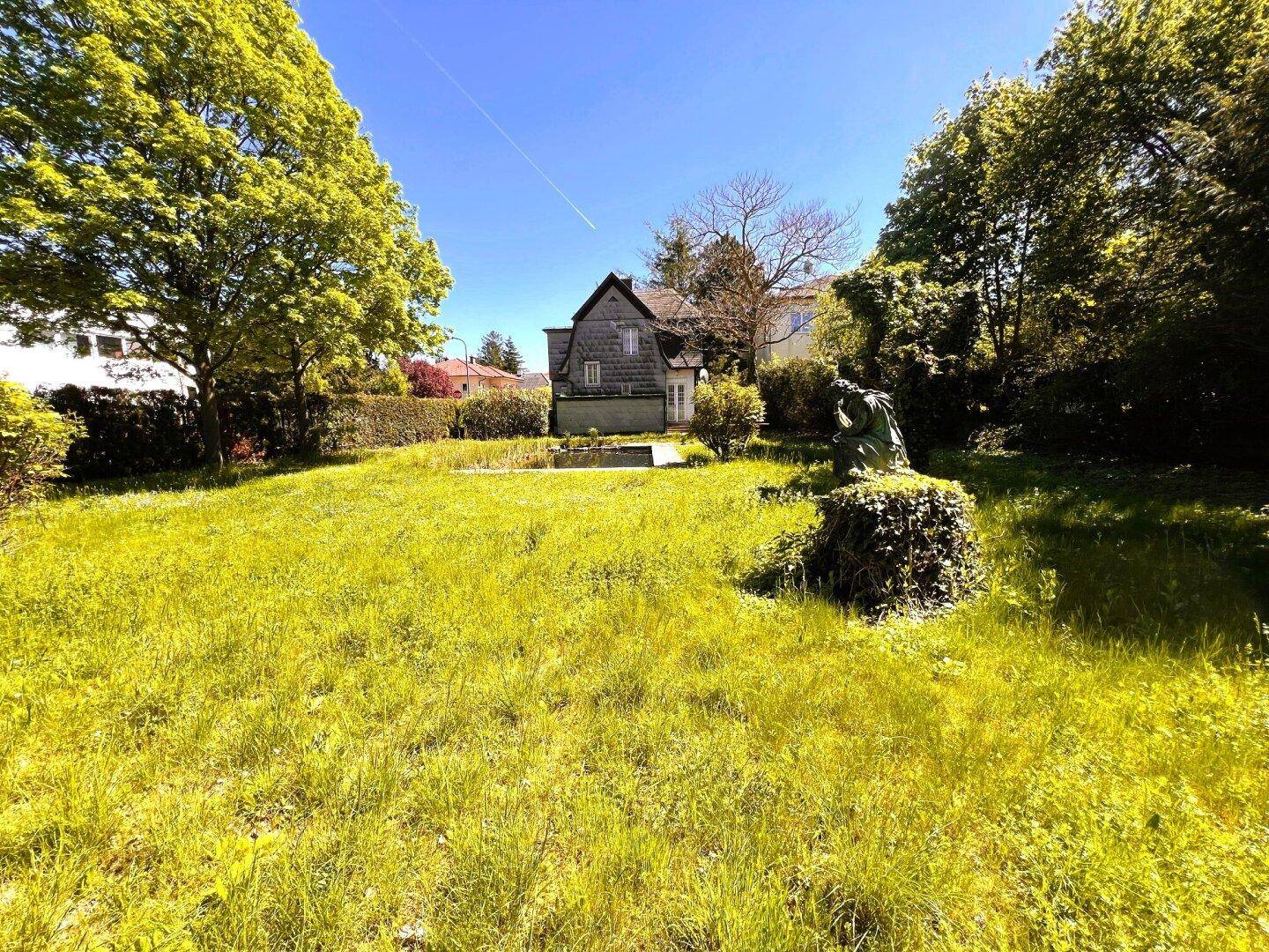 Weitläufiger Garten mit Rasenfläche und altem Baumbestand, das historische Haus ist im Hintergrund sichtbar.