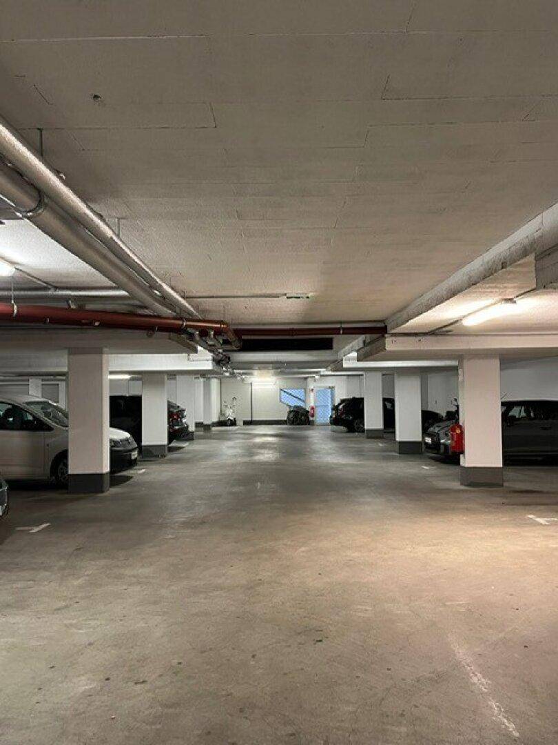 Helle Tiefgarage mit Betonboden und -säulen, ausgestattet mit Deckenbeleuchtung und sichtbaren Rohren.