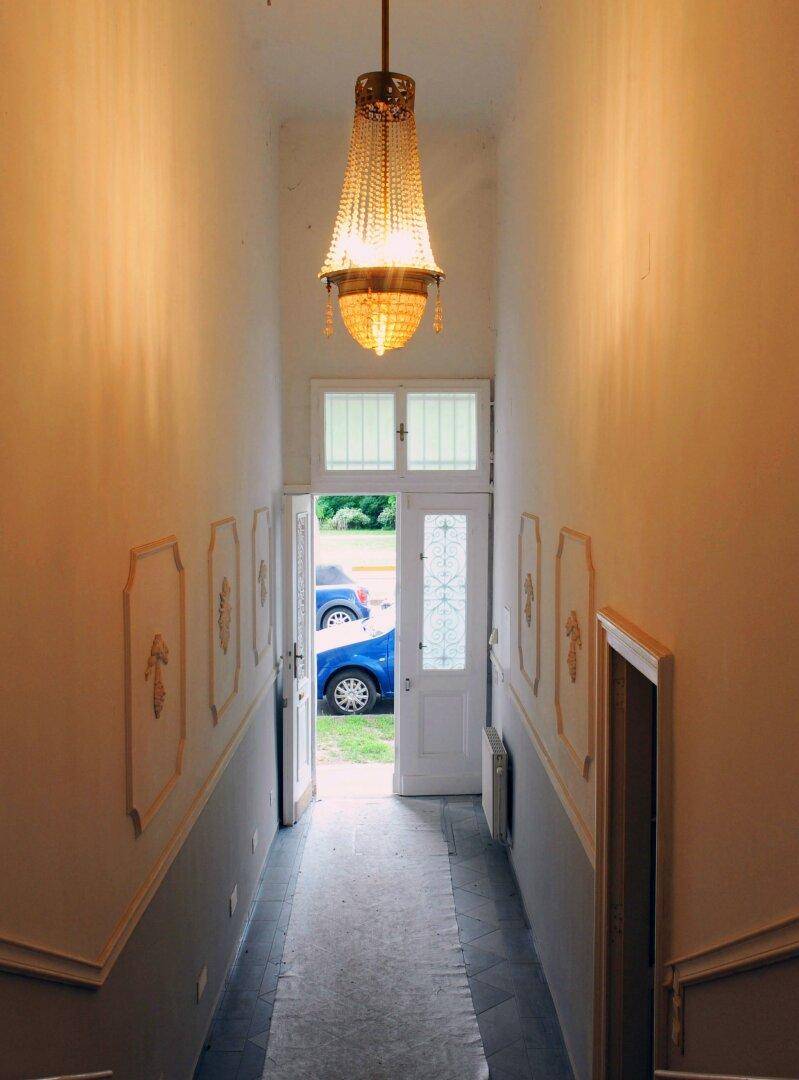 Eleganter Flur mit hoher Decke, Kronleuchter und Blick durch die Eingangstür ins Freie.