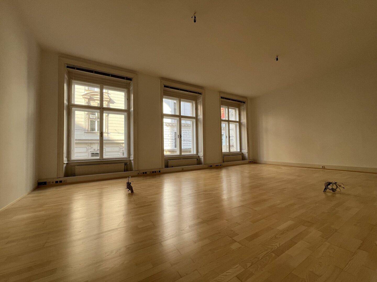 Langer, heller Raum mit drei großen Fenstern und glänzendem Holzboden.