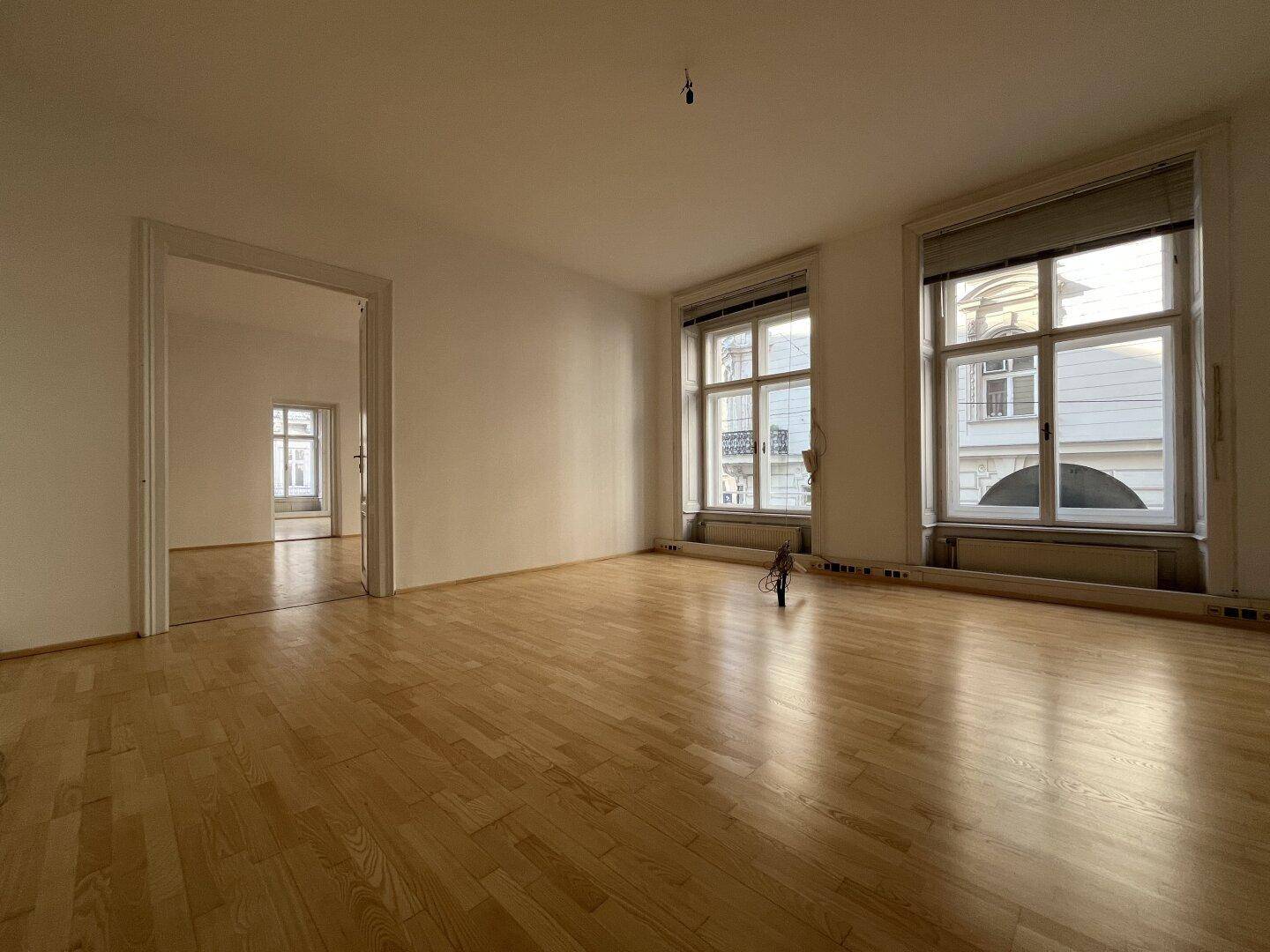Heller Wohnraum mit Holzboden und zwei großen Fenstern, die viel Tageslicht hereinlassen.