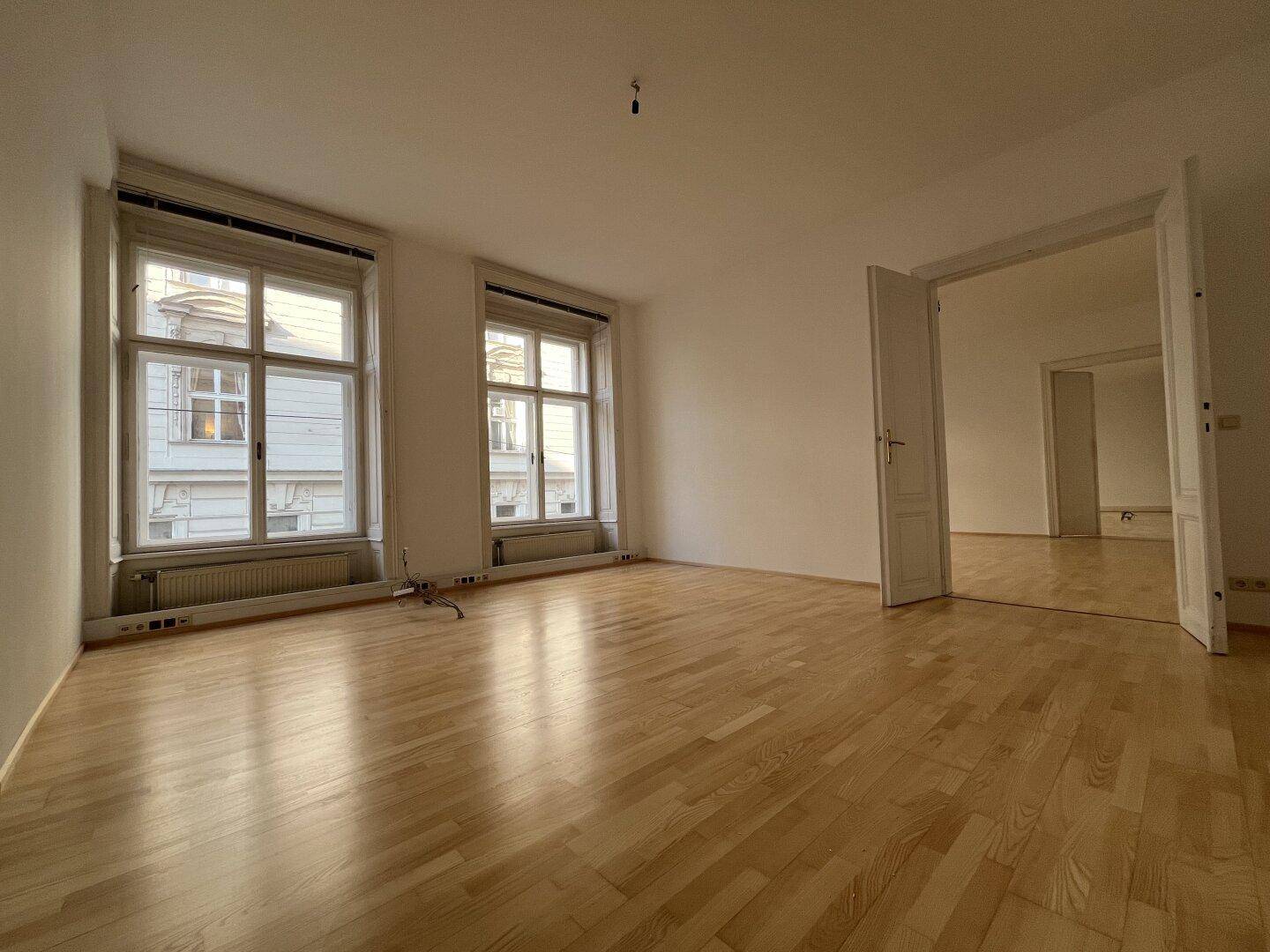 Geräumiger Raum mit Holzboden, großen Fenstern und offener Tür zu einem weiteren Zimmer.