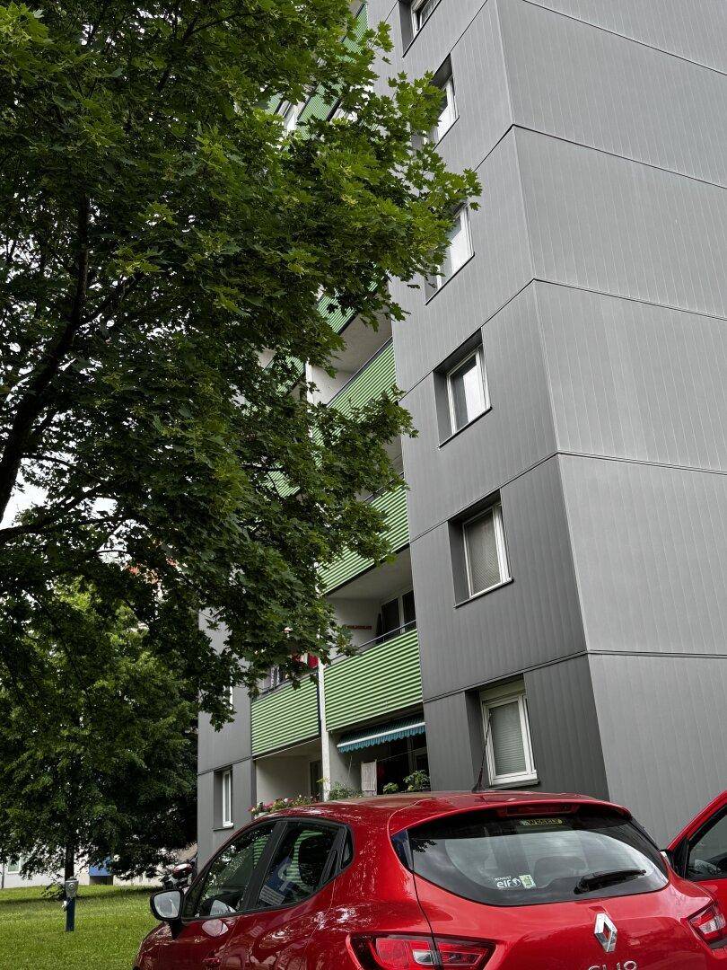 Mehrstöckiges Wohngebäude mit grauer Fassade und markanten grünen Balkonen, umgeben von Bäumen.