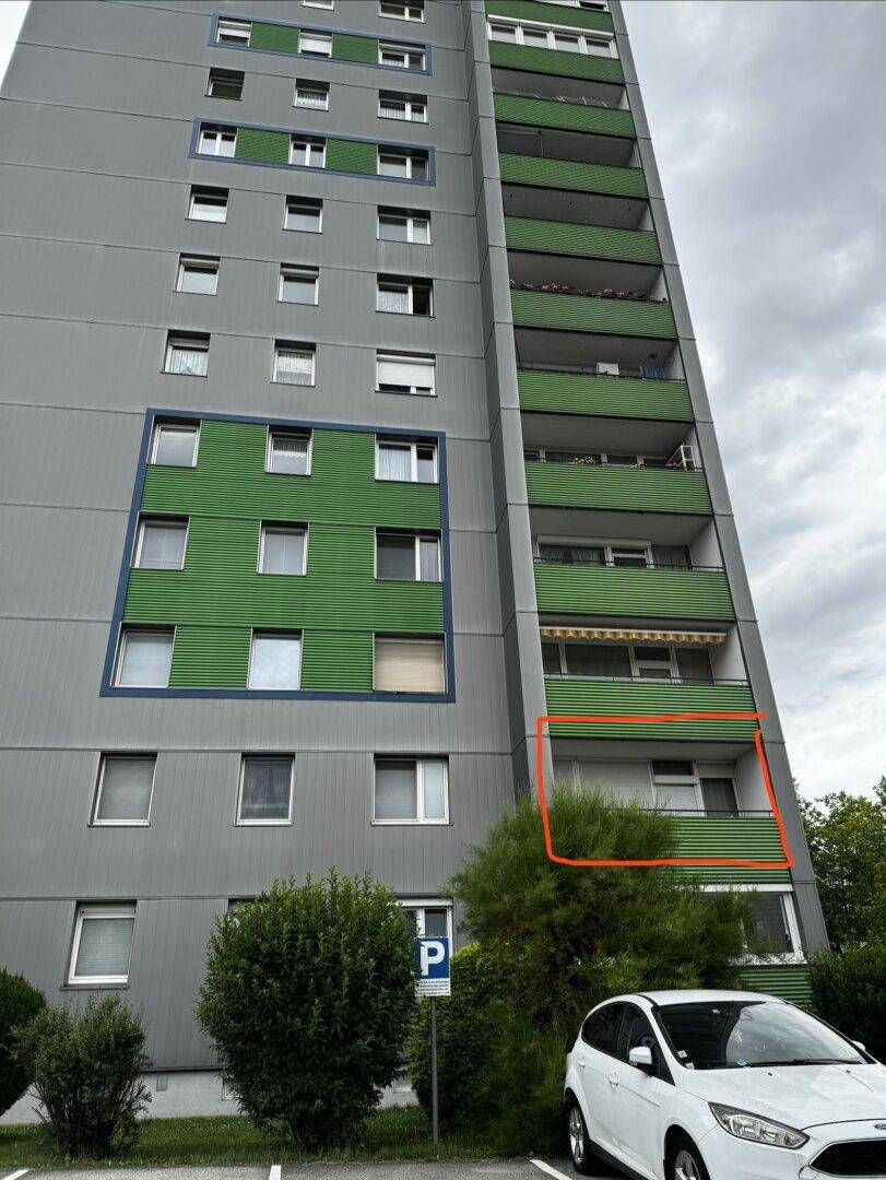 Hohes Wohngebäude mit grauer Fassade und vielen grünen Balkonen, umgeben von Grünflächen und Parkplätzen.