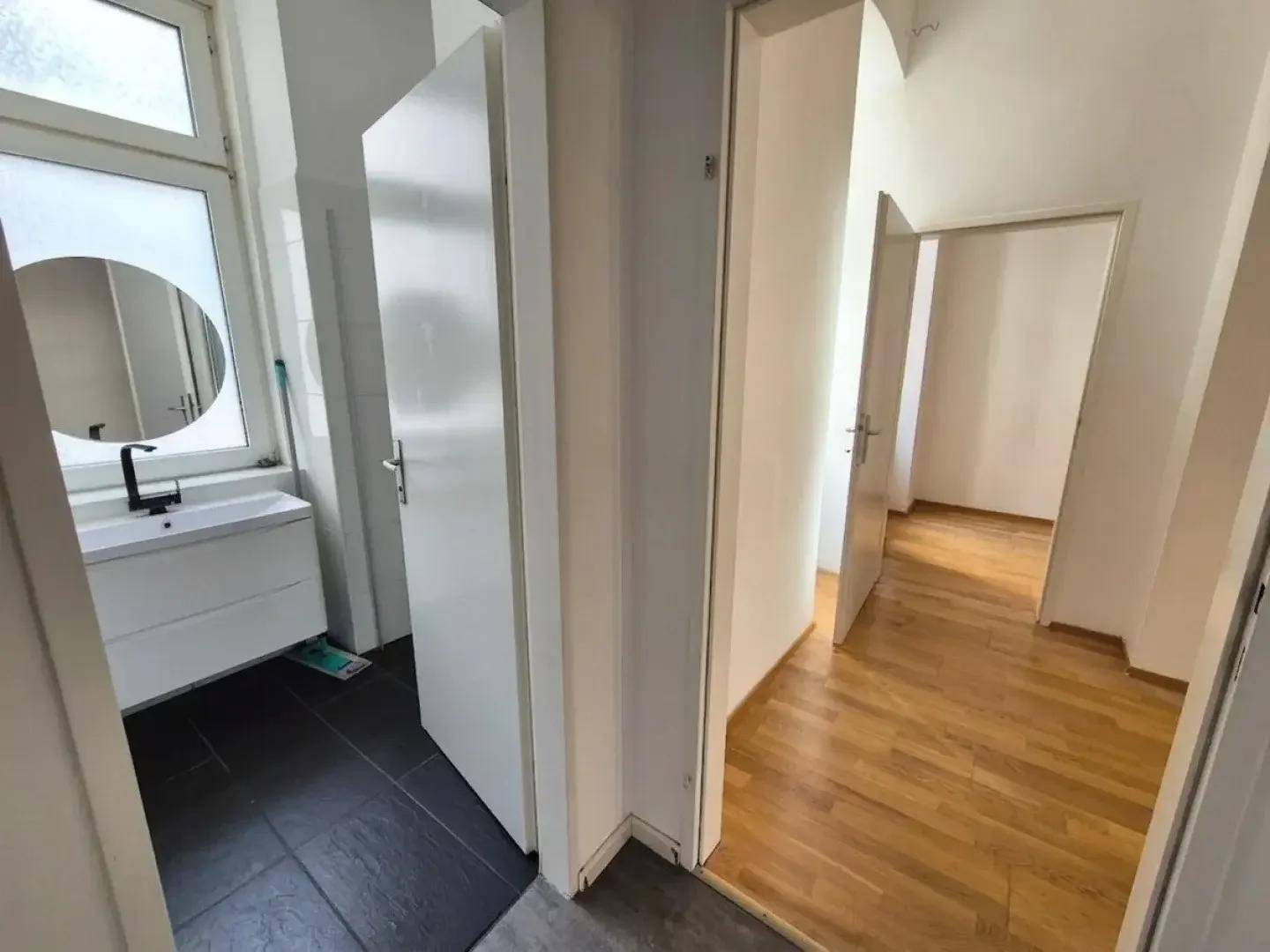 Flur mit hellem Holzparkett, der zu einem modernen Badezimmer mit dunklen Fliesen führt.