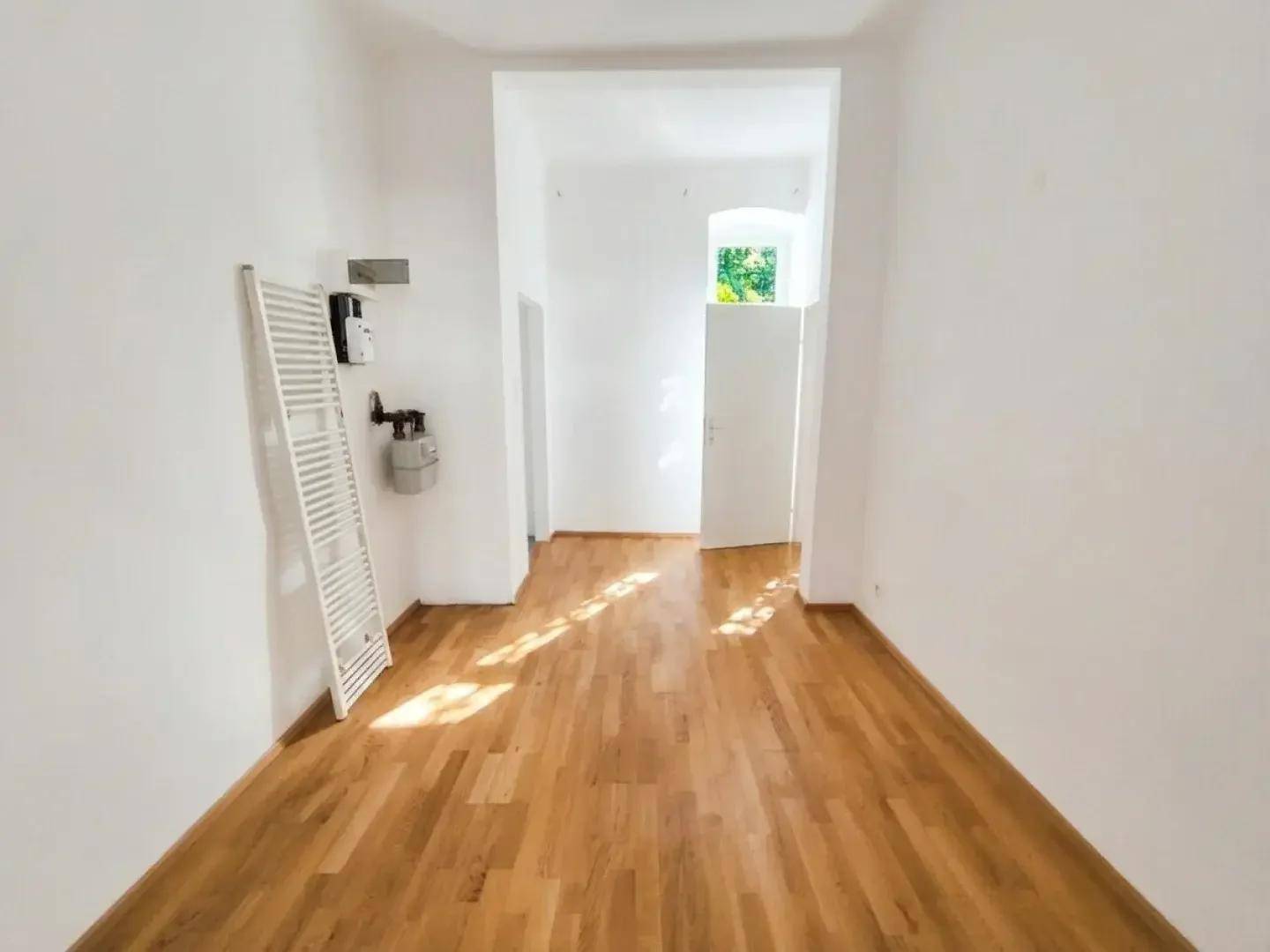 Langer, schmaler Raum mit Holzparkettboden, weißen Wänden und einem Fenster am Ende.