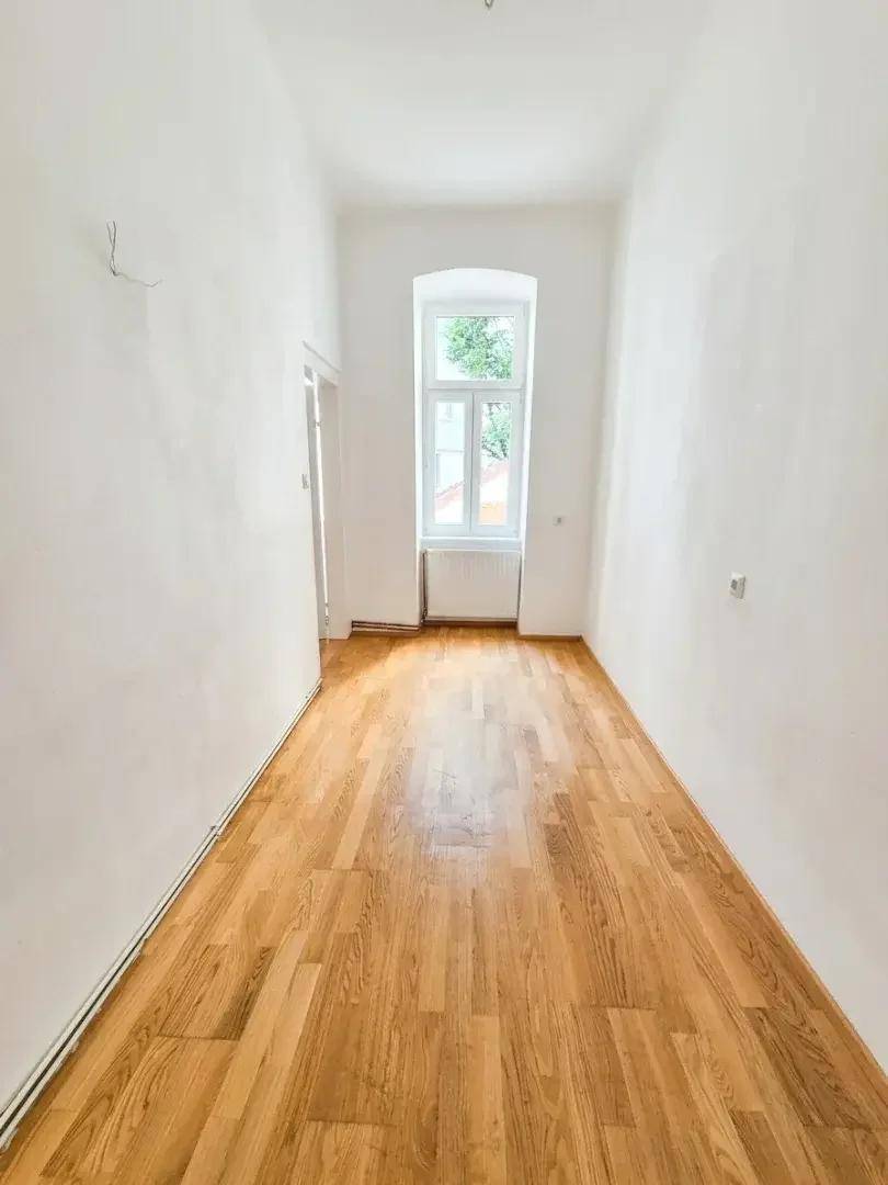 Heller, leerer Raum mit Holzparkettboden und einem hohen Fenster, das Tageslicht hereinlässt.