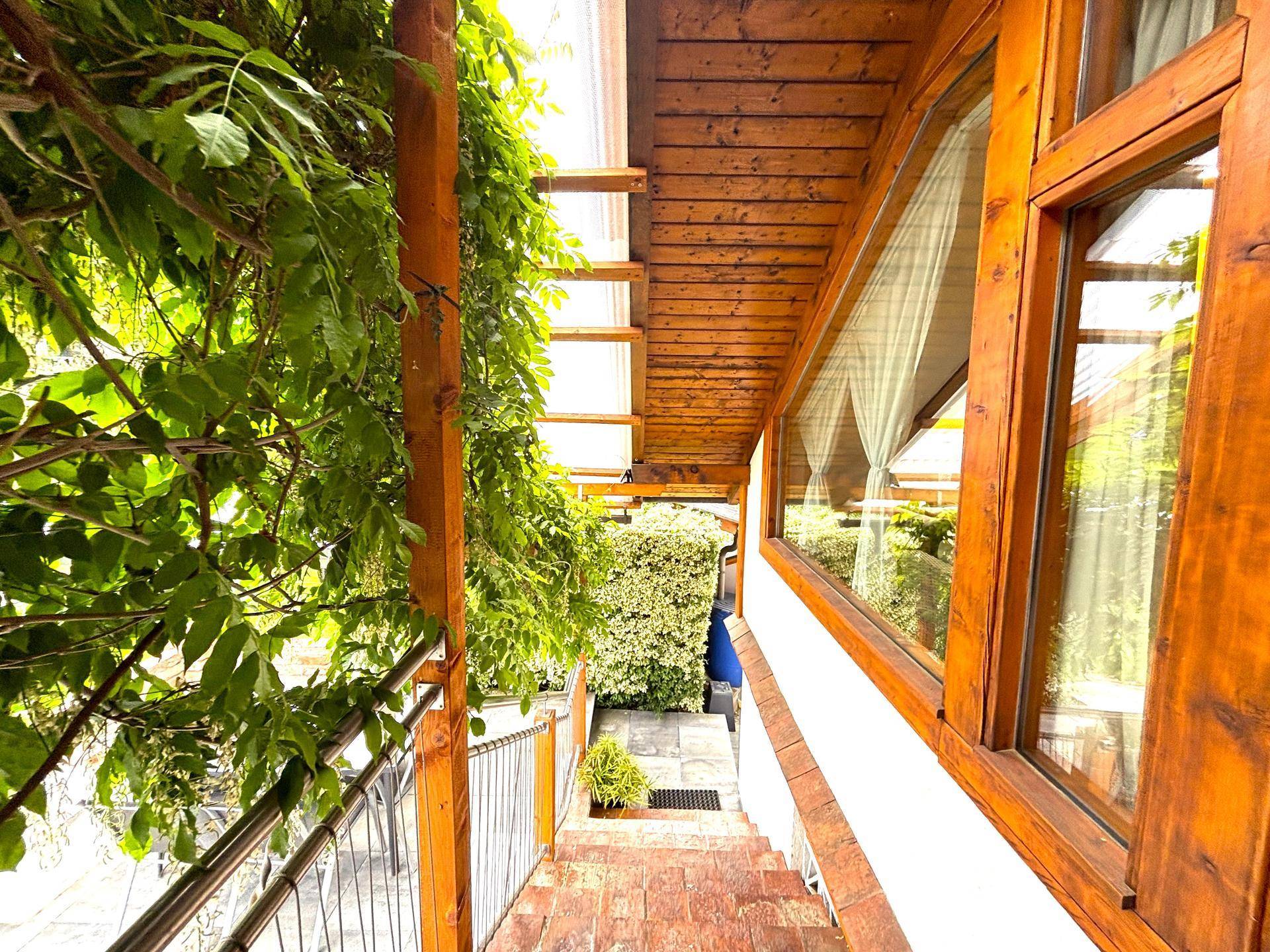 Balkon mit Holzgeländer und Treppe, umgeben von üppigem Grün und Hecken.