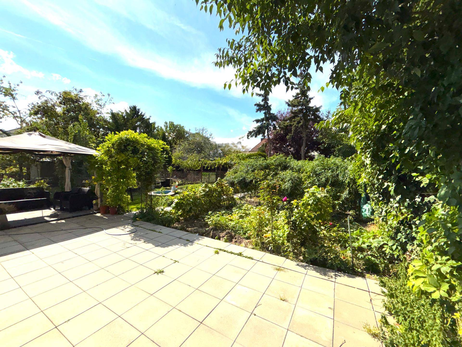 Gepflasterte Terrasse mit Gartenmöbeln unter einem Pavillon und Blick auf den üppigen Gartenbereich.