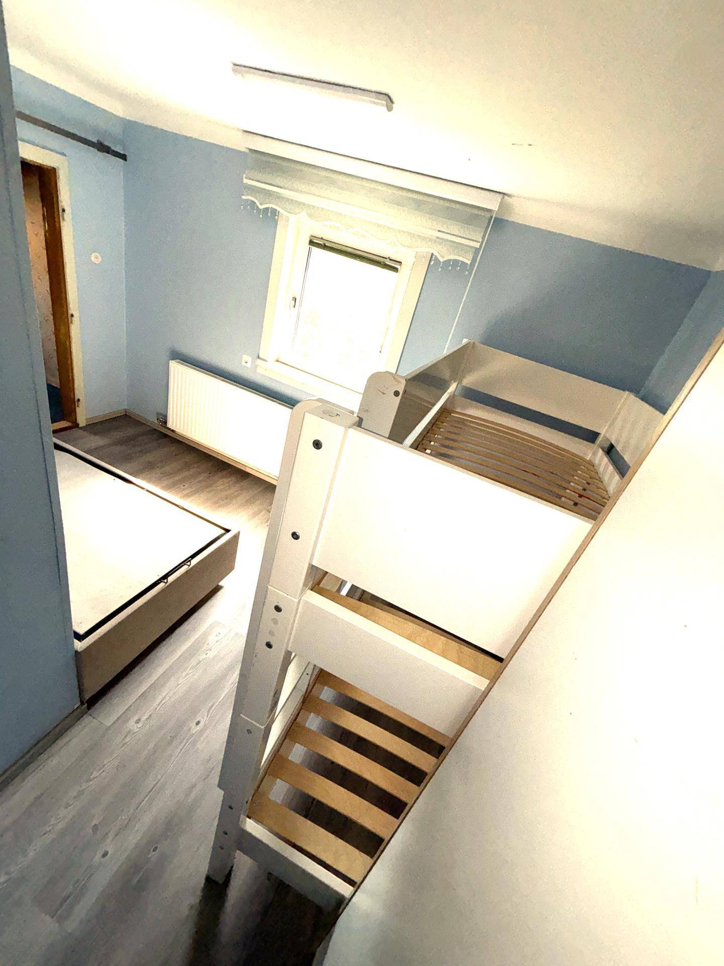 Helles Schlafzimmer mit Etagenbett, Laminatboden und Fenster mit Heizkörper unter blau gestrichenen Wänden.