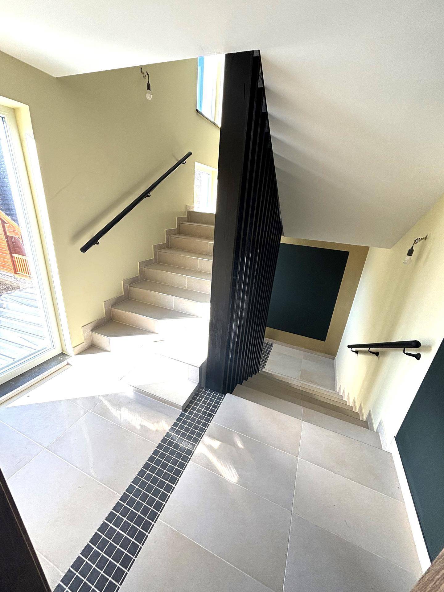 Modernes Treppenhaus mit hellen Fliesen, dunklem Geländer und viel natürlichem Licht.