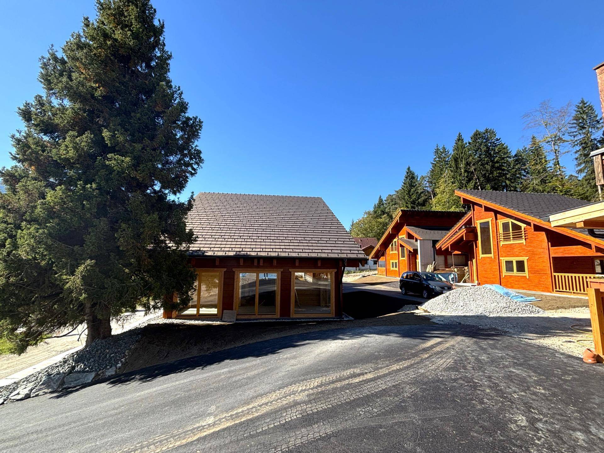 Mehrere neue Holzhäuser im Bau mit Zufahrt und umgebenden Bäumen in alpiner Umgebung.