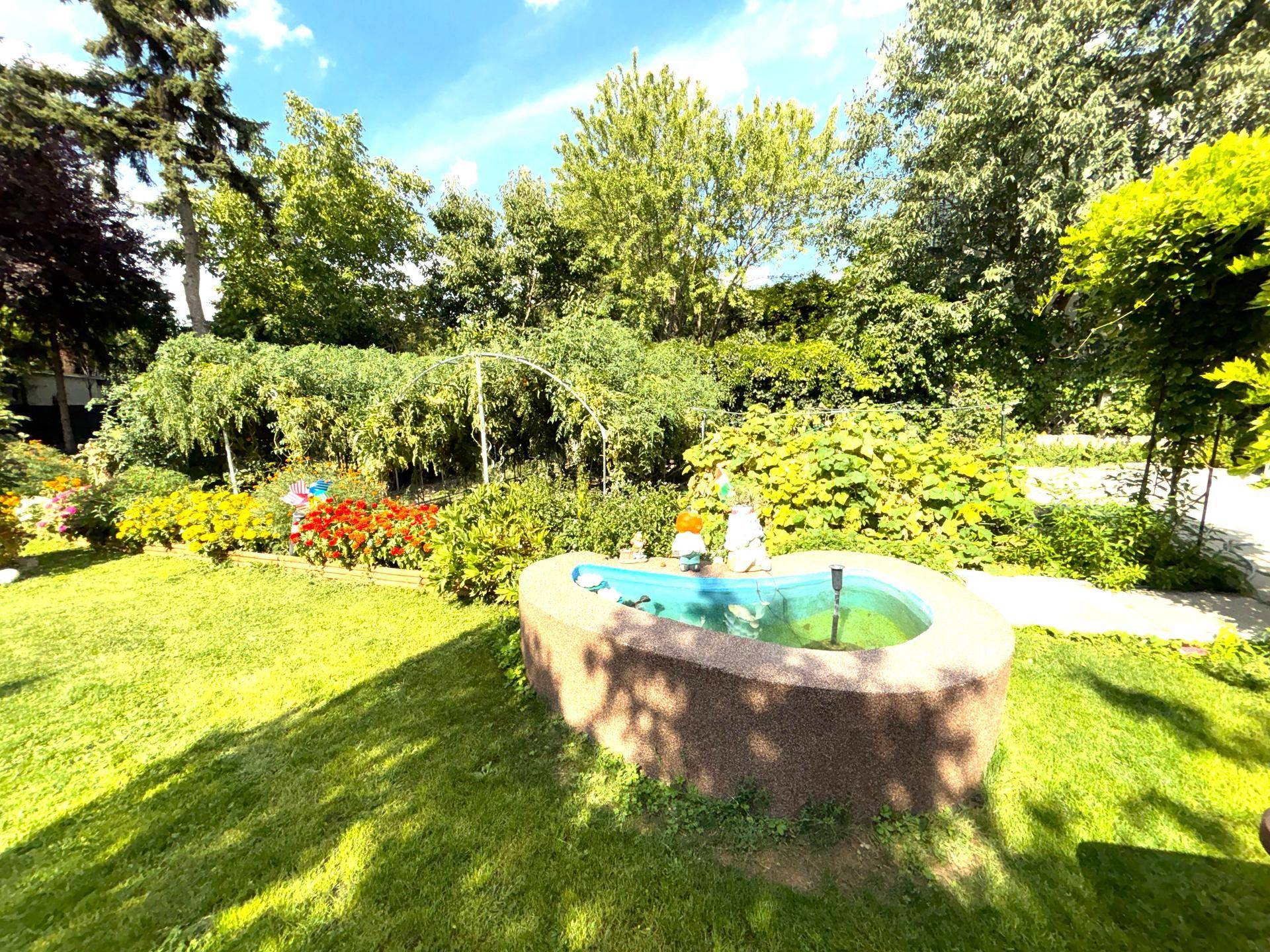 Grüner Garten mit einem dekorativen Brunnen, bunten Blumenbeeten und Gartenfiguren unter blauem Himmel.