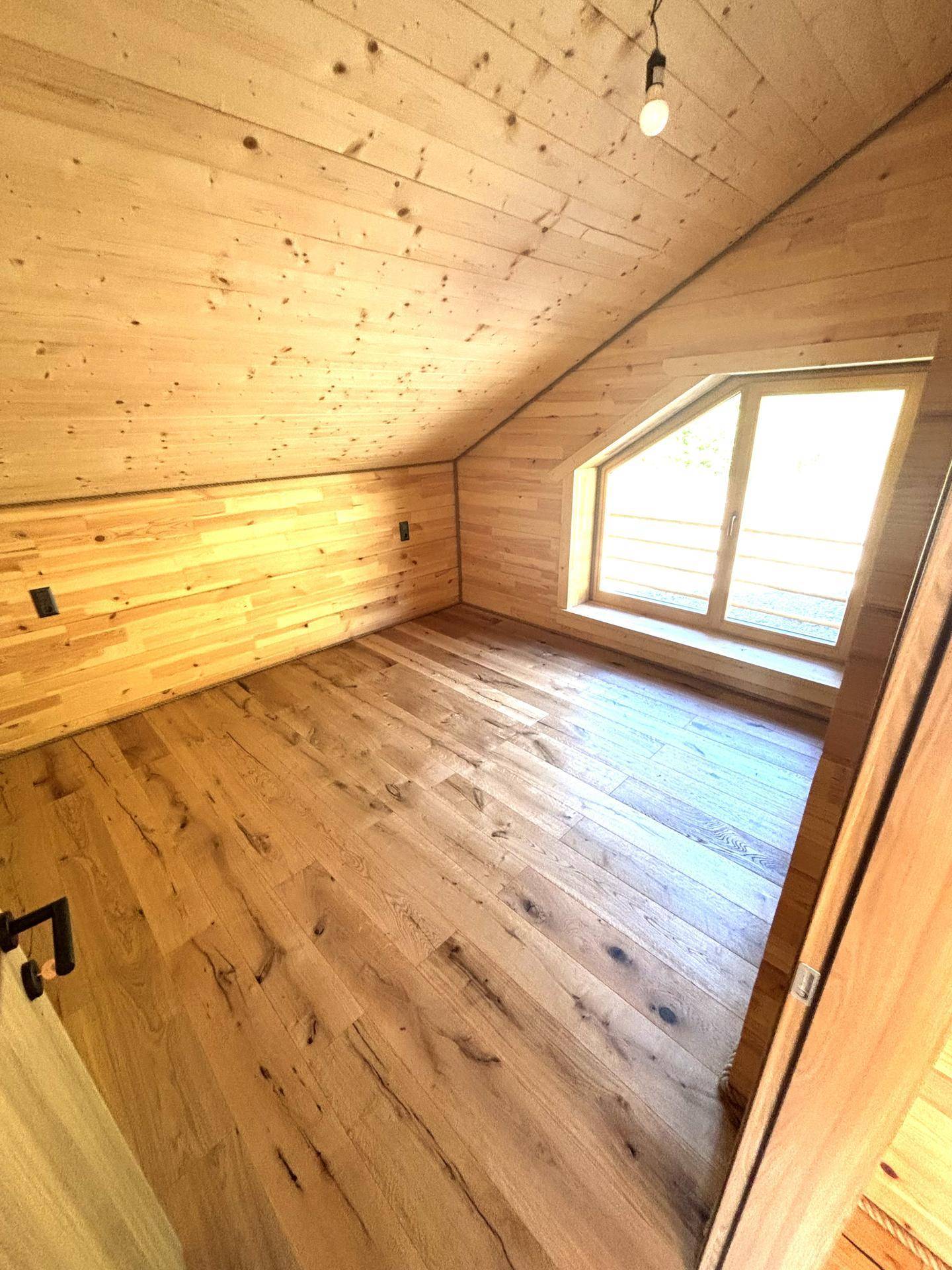 Helles Schlafzimmer im Dachgeschoss mit Holzboden, Holzvertäfelung und schrägem Fenster.