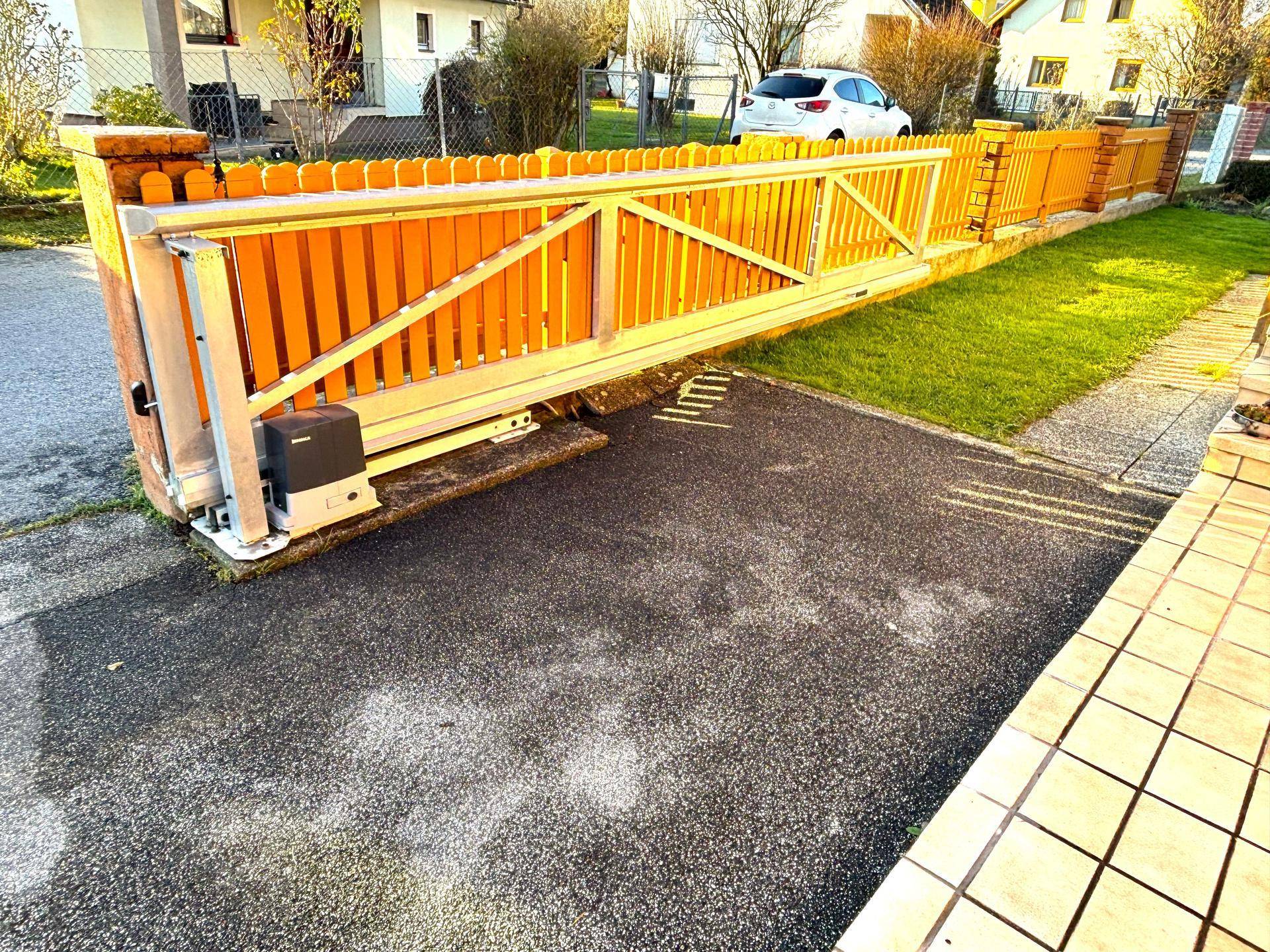 Automatisches Schiebetor aus Holz mit gelbem Anstrich und asphaltierter Einfahrt zum Grundstück.
